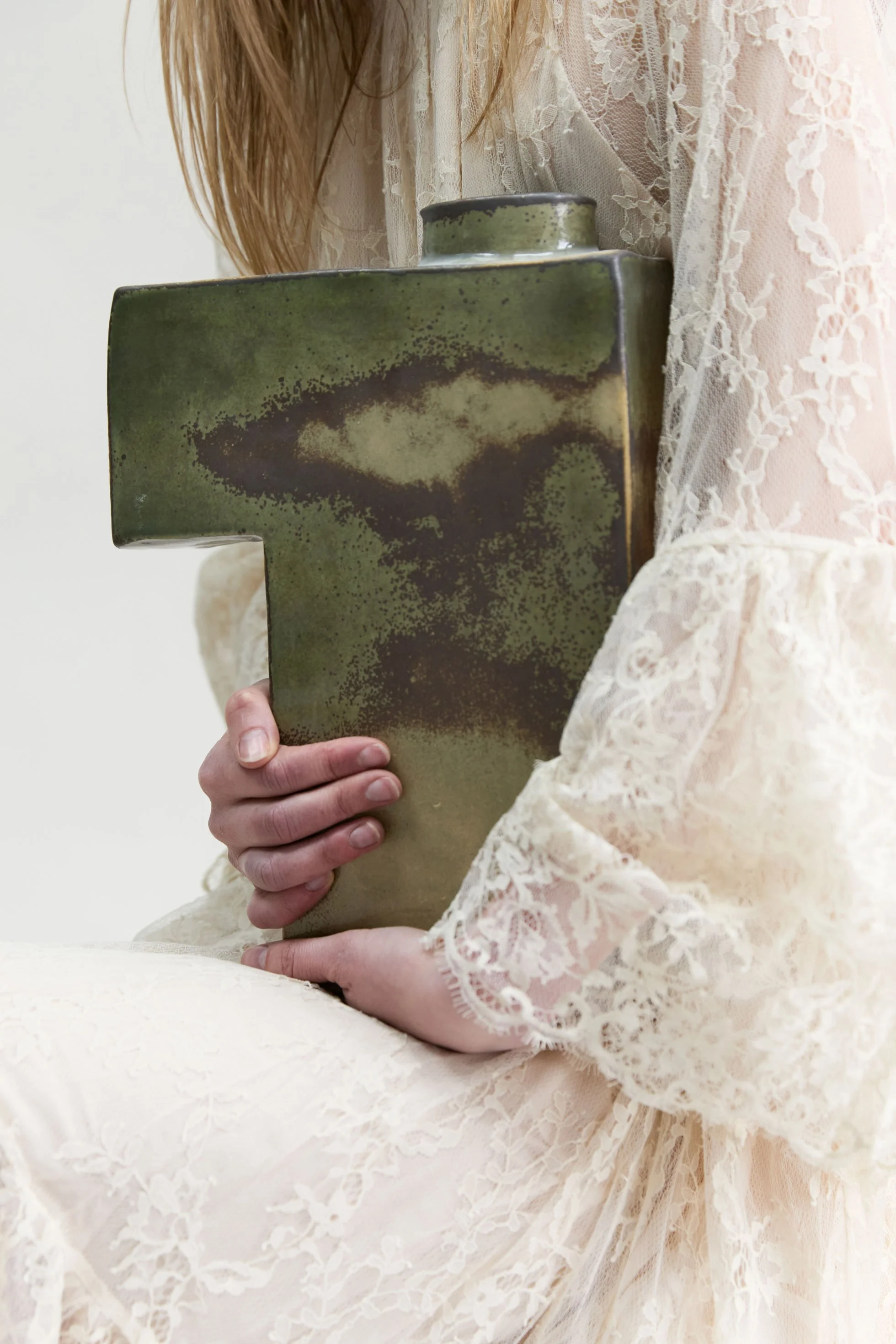 Une femme en robe en dentelle beige ou blanche tient une croix en céramique verte.