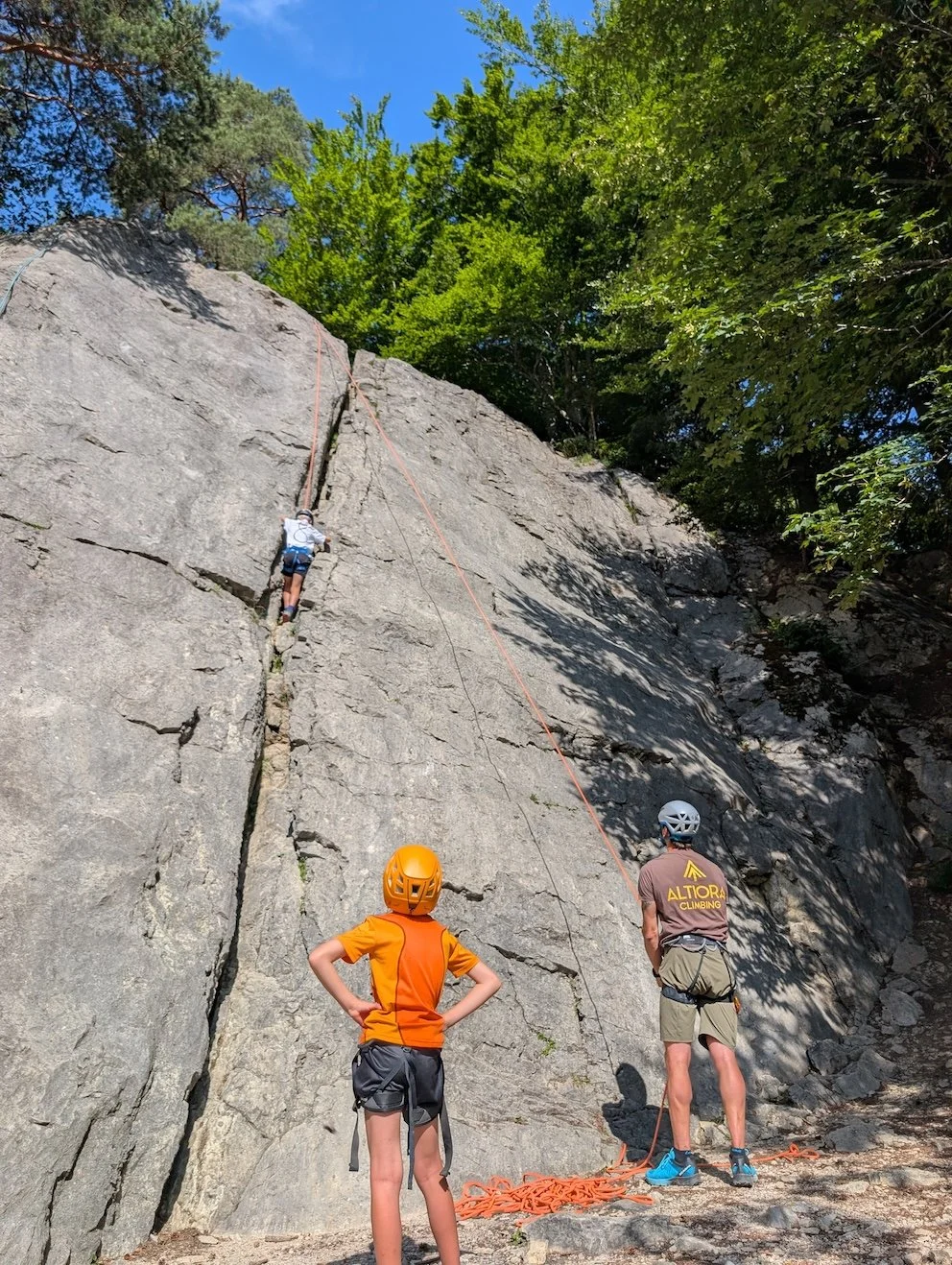 A climbing instructor explaining safety procedures to two climbers on a rock face outdoors, all wearing helmets and harnesses.