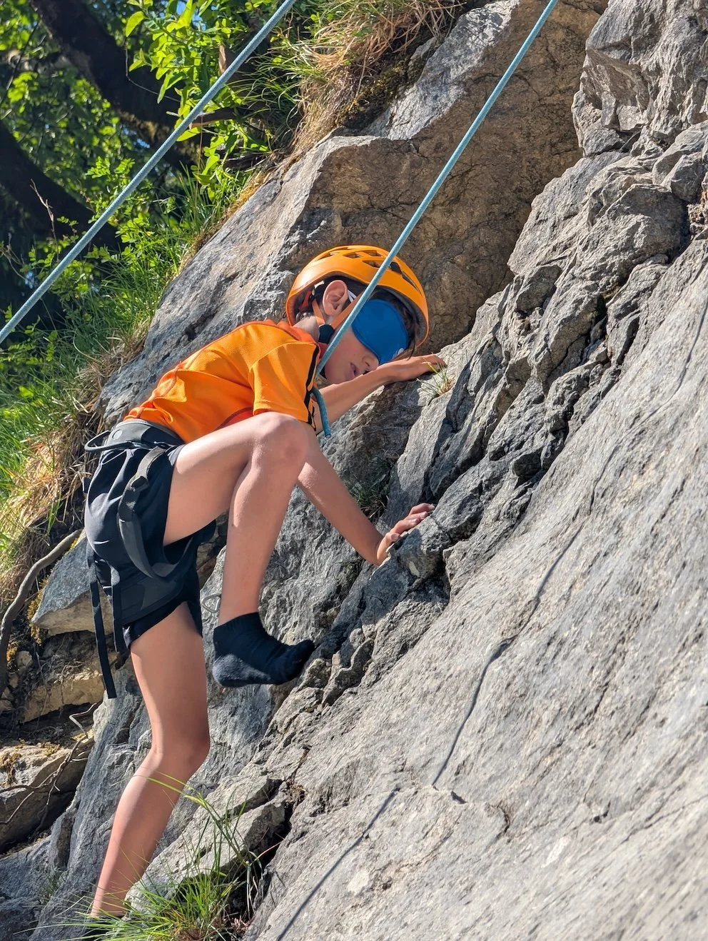 A woman rock climbing wearing a white helmet and black shirt, secured with a harness, climbing a rock face, with another person below on the ground assisting, surrounded by trees and nature.