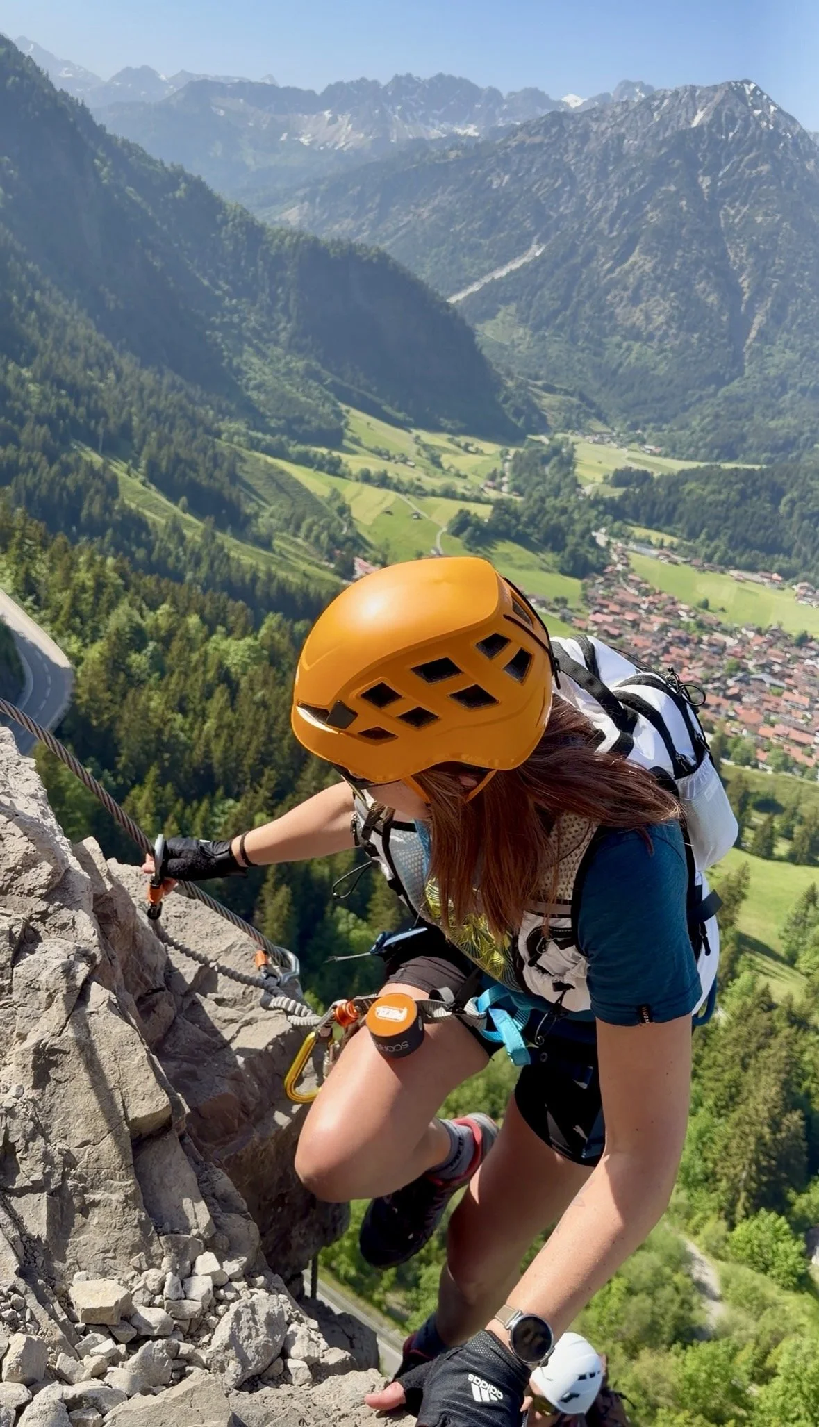 A person rock climbing on a vertical rock face with climbing gear, helmet, and harness, while another person stands below with climbing equipment.