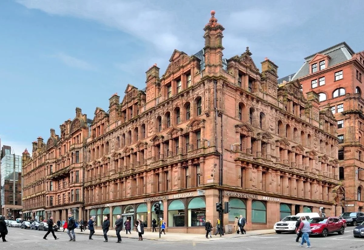 A historic red sandstone building with ornate architectural details at street level, with pedestrians walking in front and cars passing by, under a partly cloudy sky.