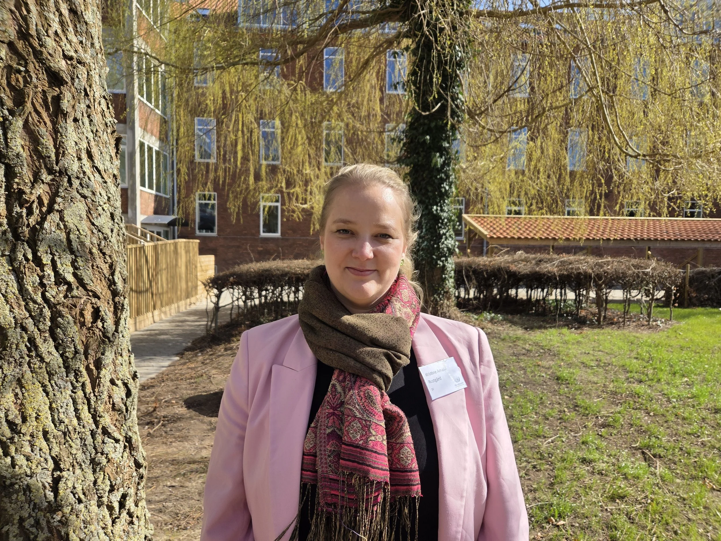 Kvindelig person med lang, blond hår, iført sort top og grøn jakke, smilende foran hvid baggrund.