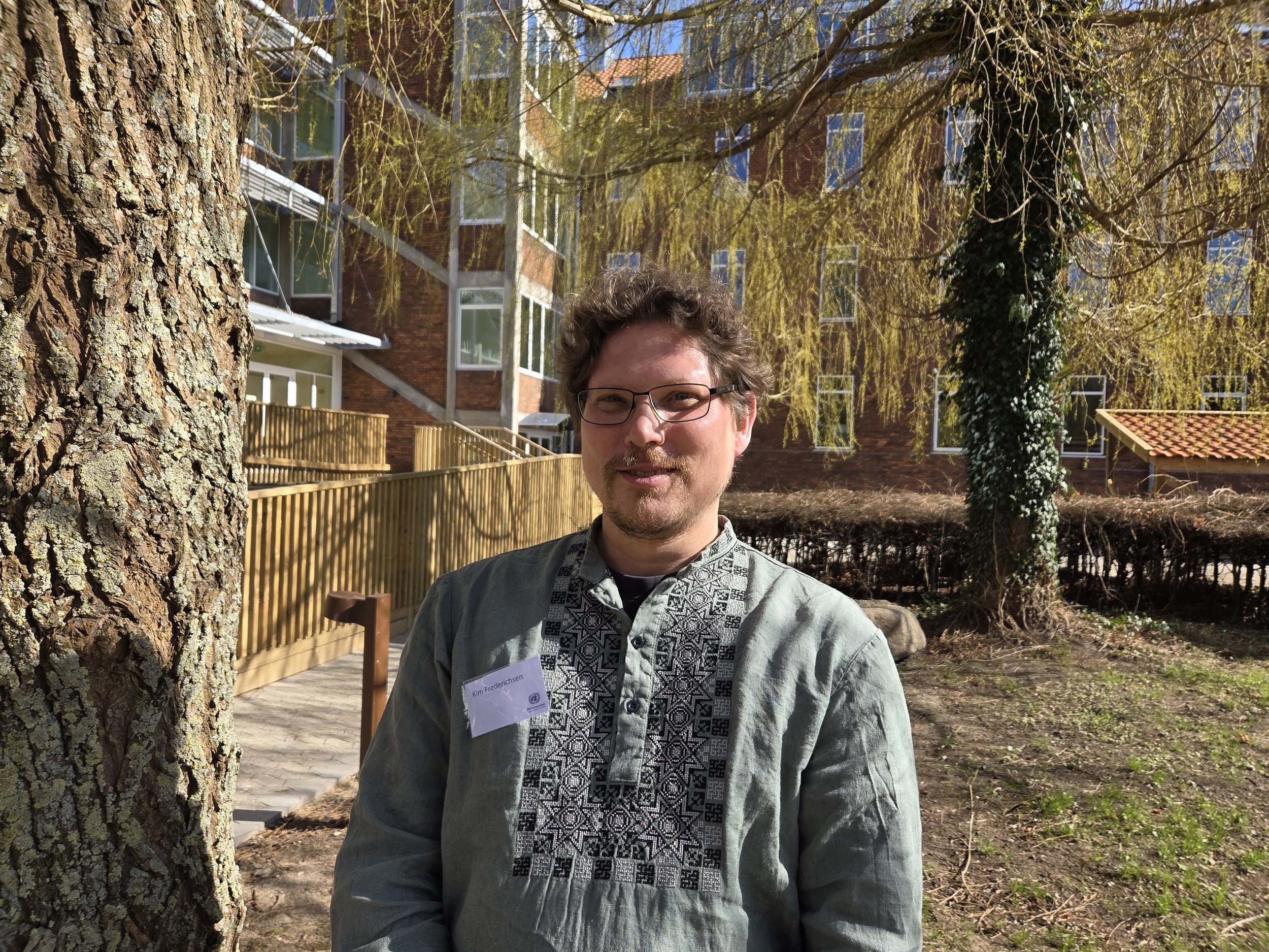 En ung mand med briller, let skæg og vasket hår, iført en sort sweatshirt over en rutig skjorte, smilende foran en hvid væg.