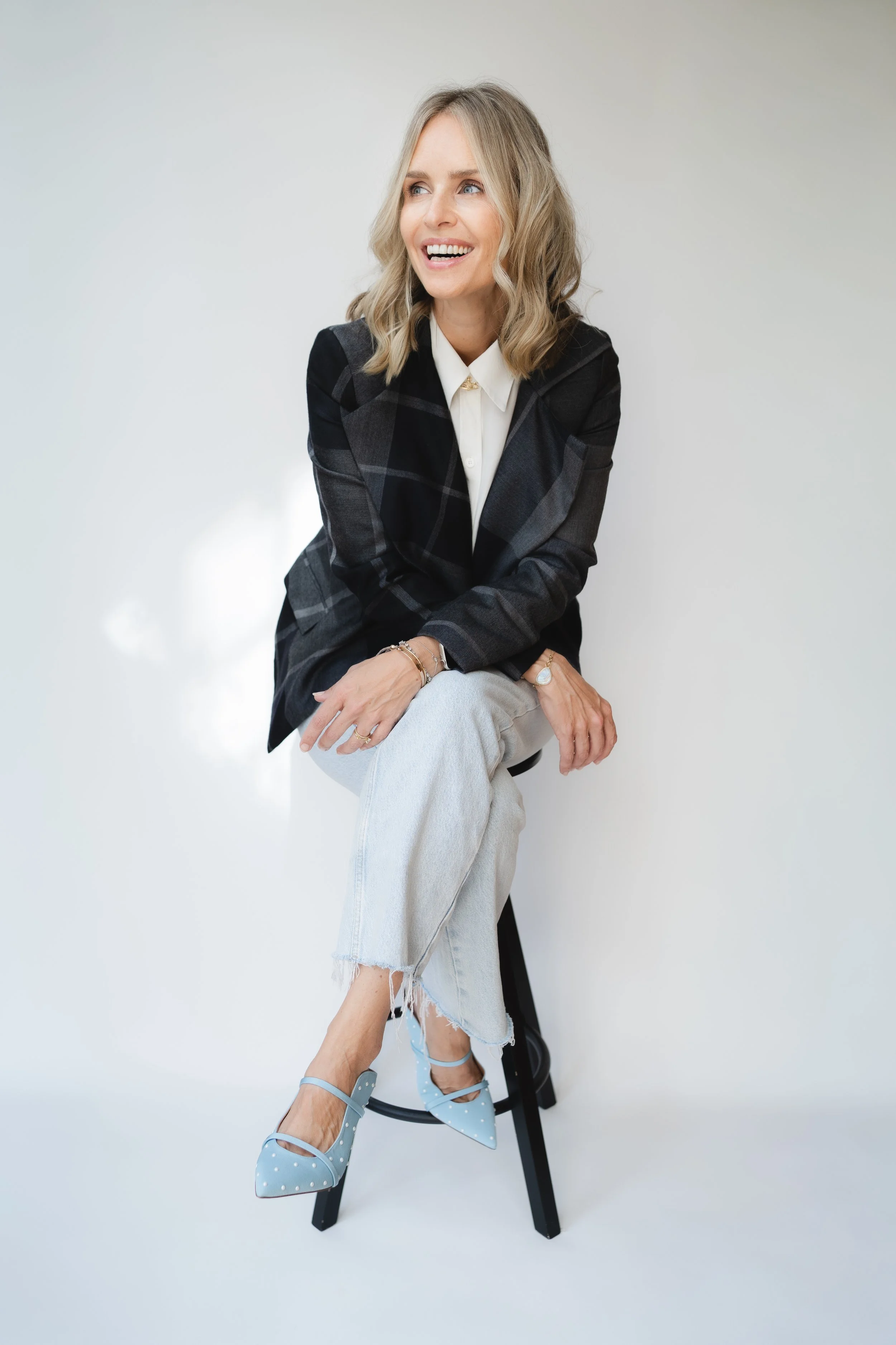 A woman with blonde wavy hair wearing a dark plaid blazer, white shirt, light-colored ripped jeans, and blue polka dot high heels, sitting on a black stool against a plain white background, smiling and looking to her left.