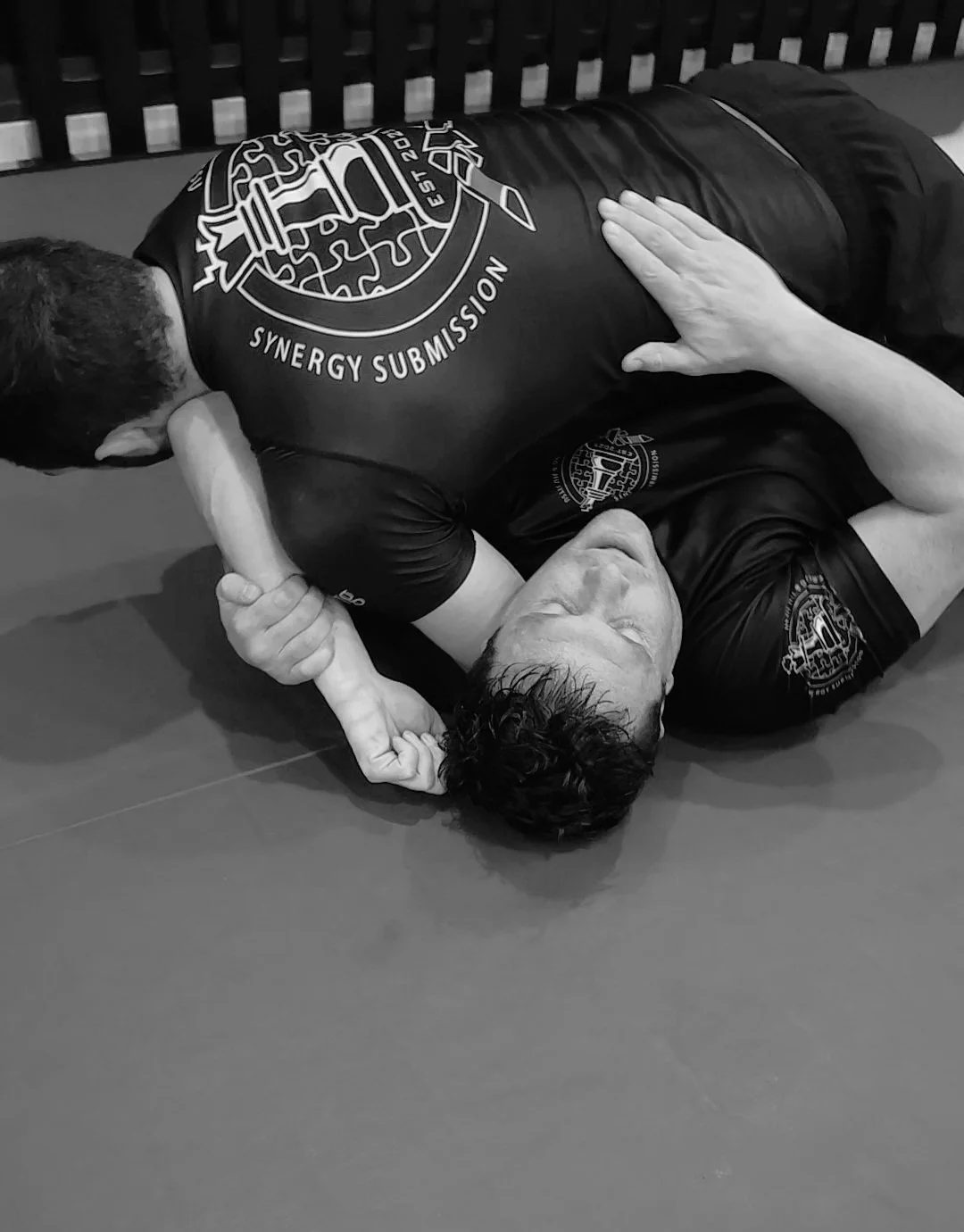 Two people practicing Brazilian Jiu-Jitsu on a mat, wearing matching shirts with a logo that reads "Synergy Submission".