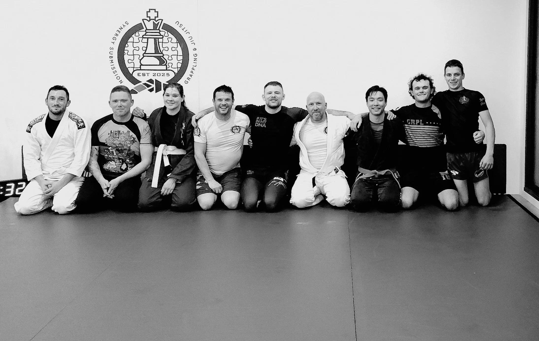 Group of people in Brazilian Jiu-Jitsu gis and casual clothing kneeling together in a gym, smiling with arms around each other, with a logo on the wall behind them.