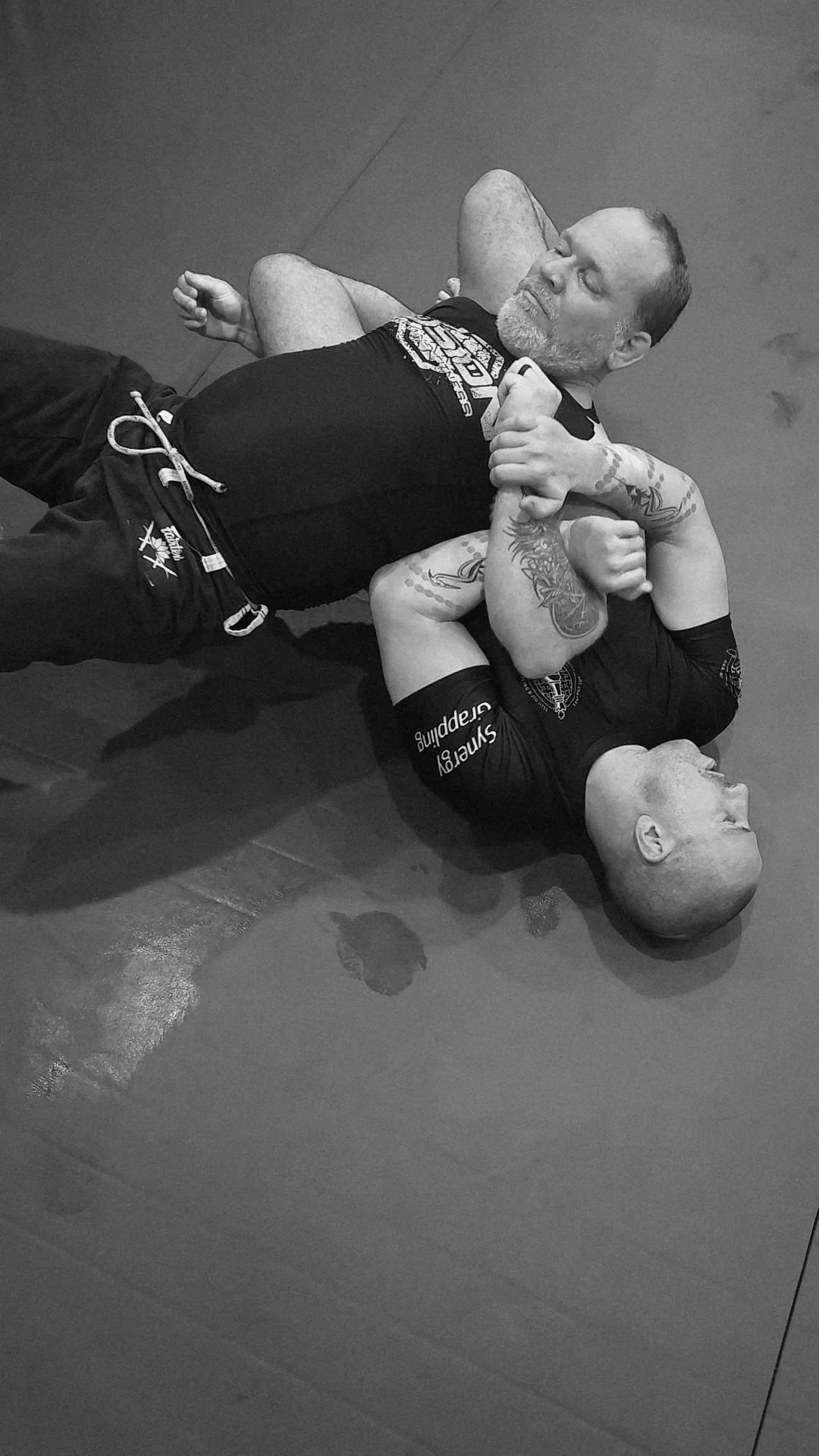 Two men practicing Brazilian Jiu-Jitsu on the floor, one in a dominant position with a tattooed arm, wearing a black rash guard, and the other in a vulnerable position with their head on the mat, shirtless with tattoos.