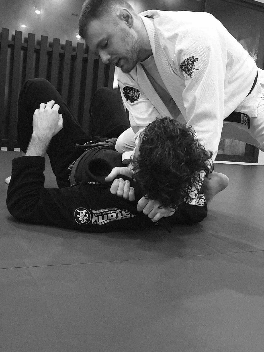 A person in a white Jiu-Jitsu gi demonstrating a chokehold on another person in a black gi, both lying on a mat, with the instructor applying pressure and the student attempting to defend.