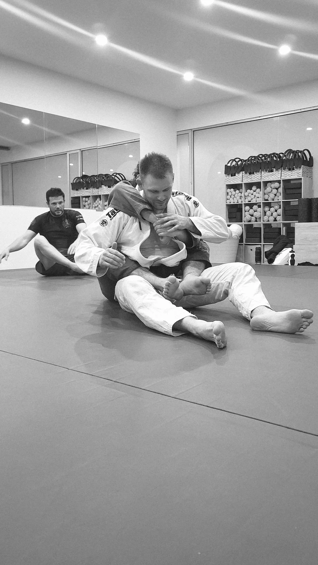 Two men practicing Brazilian Jiu-Jitsu on a mat, with one applying a choke hold on the other, in a training facility with shelves of gym equipment in the background.