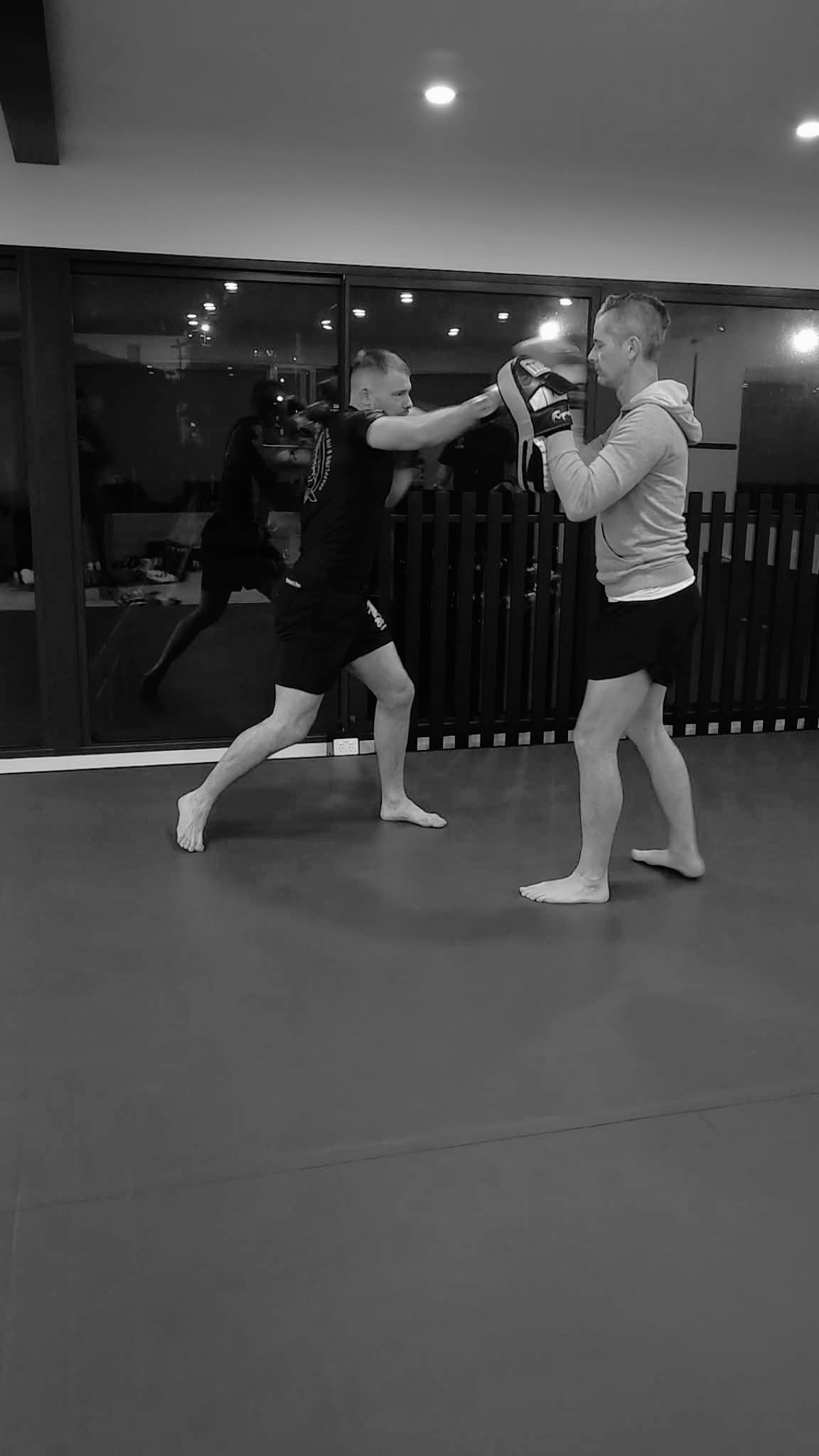 Two men practicing martial arts or boxing in a gym, with one holding focus mitts and the other performing a punch. Both are barefoot and in athletic clothing.
