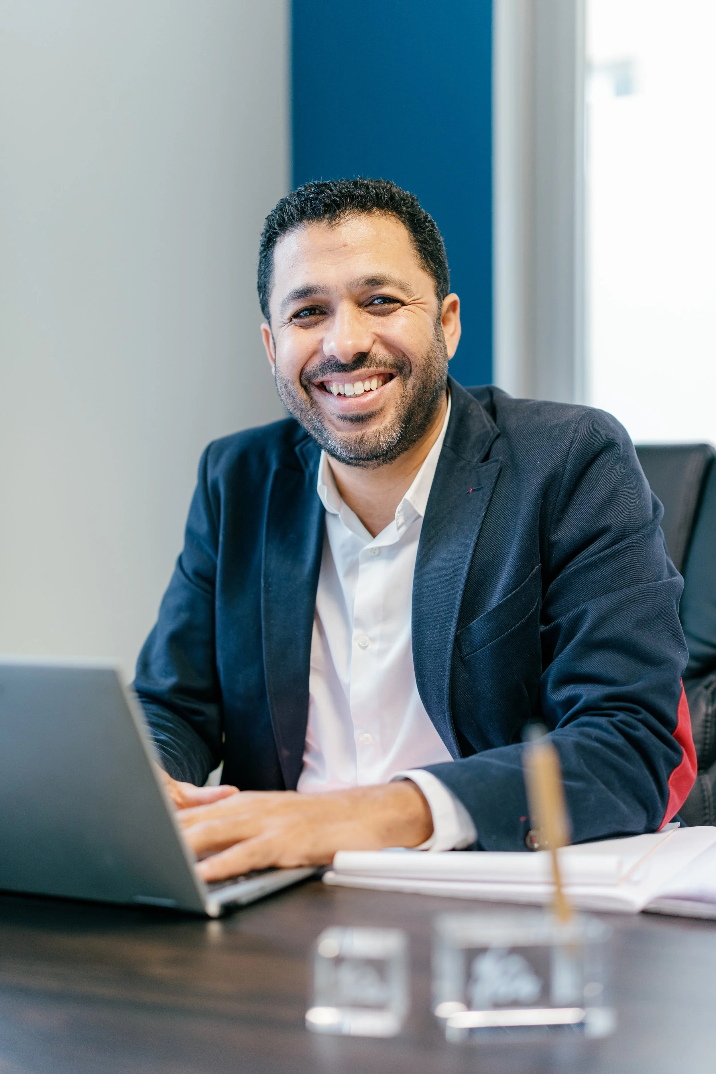 Homme souriant en costume assis à un bureau avec un ordinateur portable dans un bureau moderne.