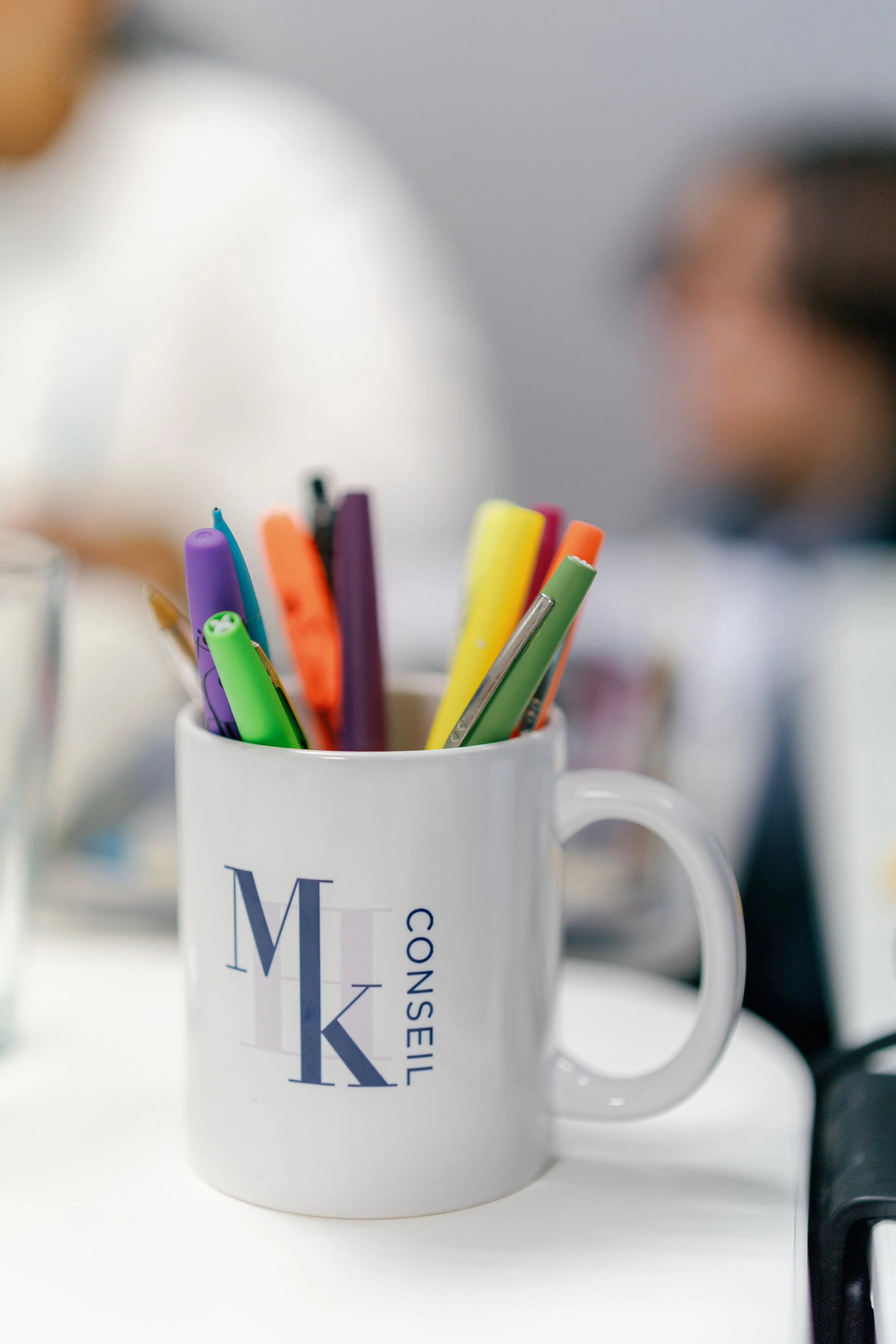 Une tasse blanche avec des stylos de différentes couleurs, portant le logo MK Conseil.