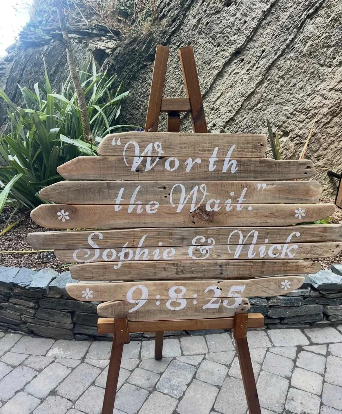 A wooden sign on an easel with the wedding date, names, and a quote, placed outdoors on a brick pathway near plants and a stone wall.