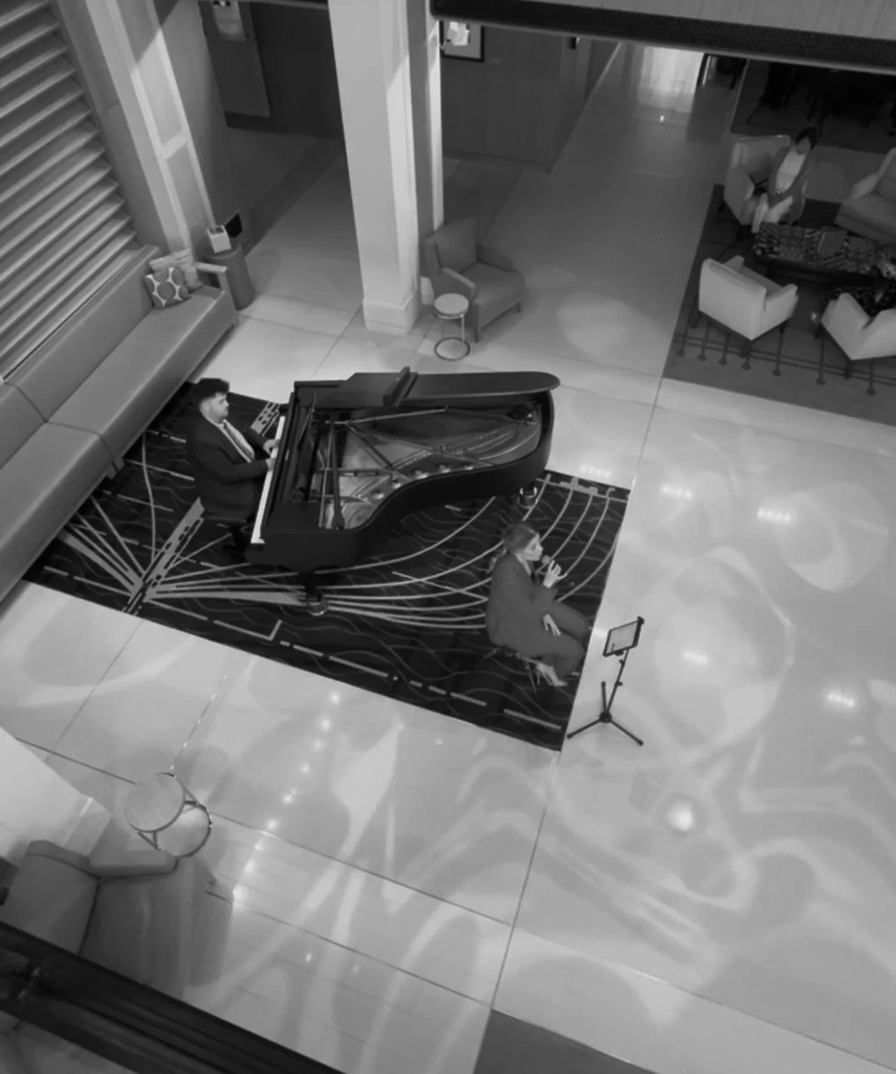 A person playing a grand piano on a rug, with another person sitting nearby speaking into a microphone, in a lobby area with seating and decorations.