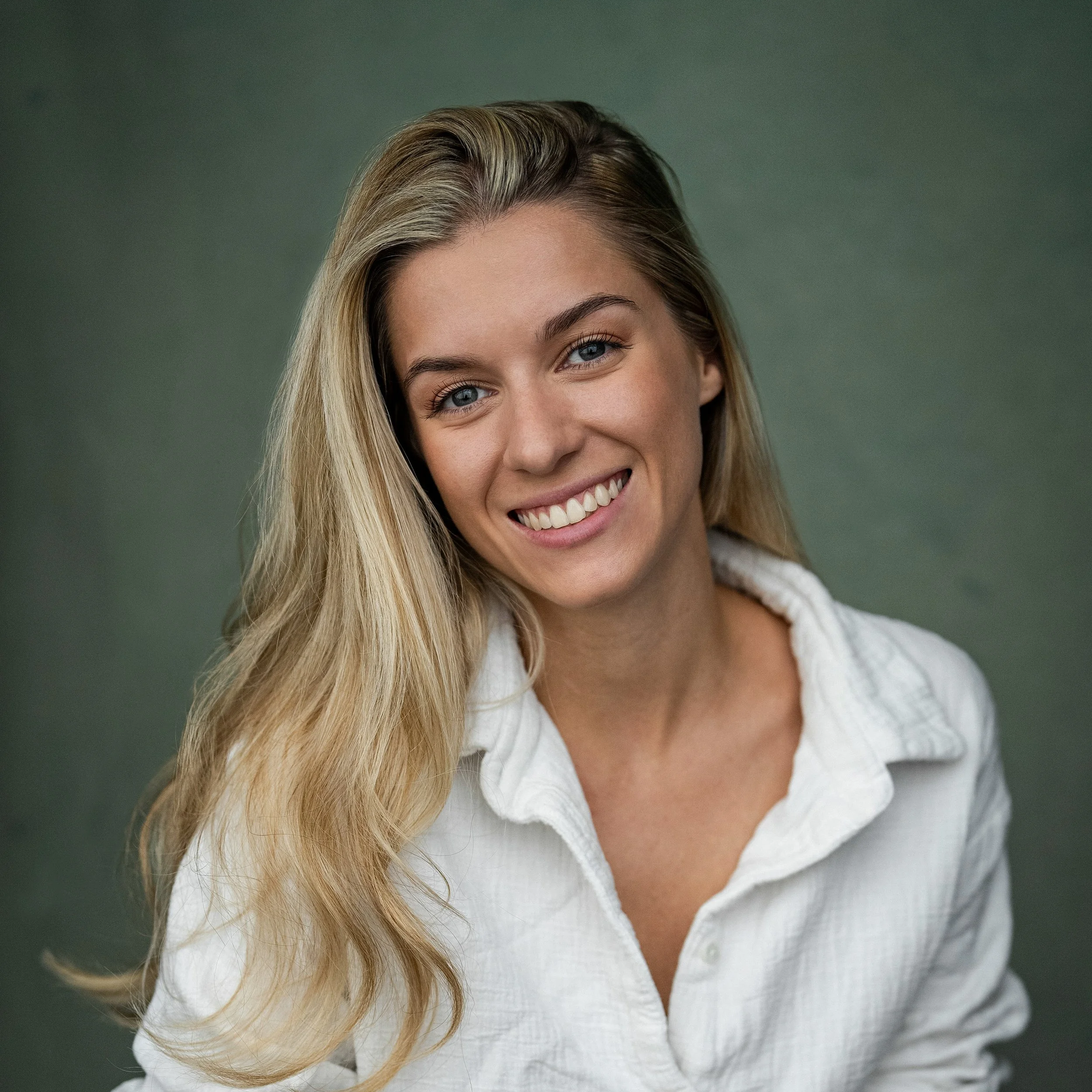 Portrait of a young woman with long blonde hair and blue eyes smiling at the camera, wearing a white shirt, with a green background.