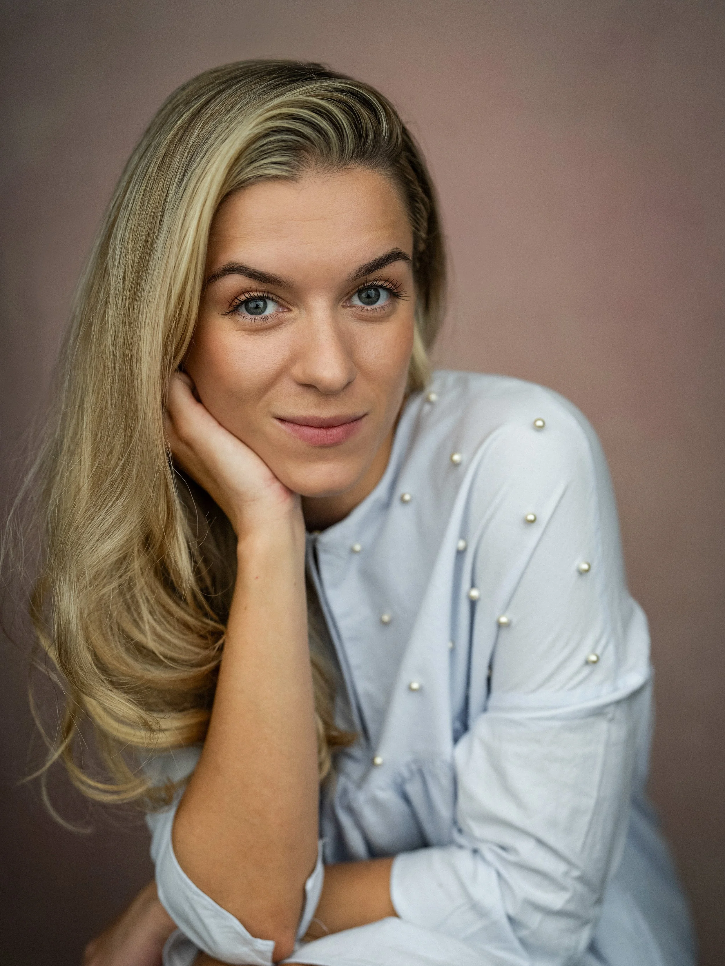 A woman with long blonde hair and blue eyes resting her chin on her hand, wearing a white shirt with pearl embellishments, looking at the camera with a slight smile.