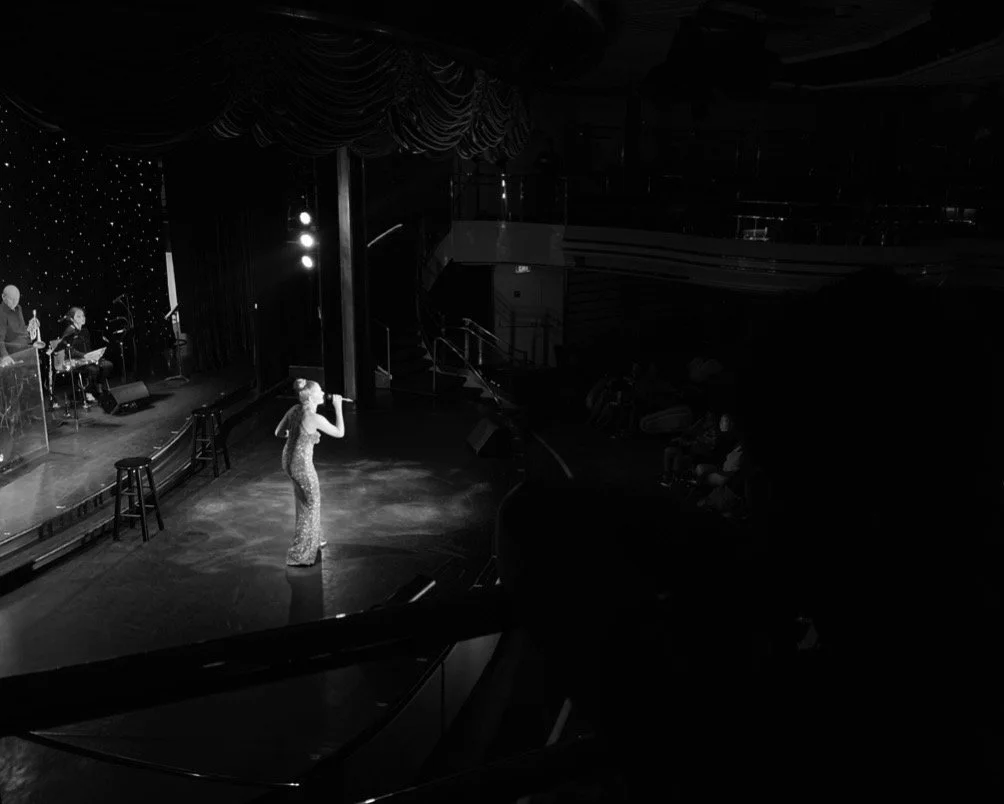 A woman in a sparkly dress performing or singing on stage with musicians playing instruments behind her, in a dark venue with spotlights.