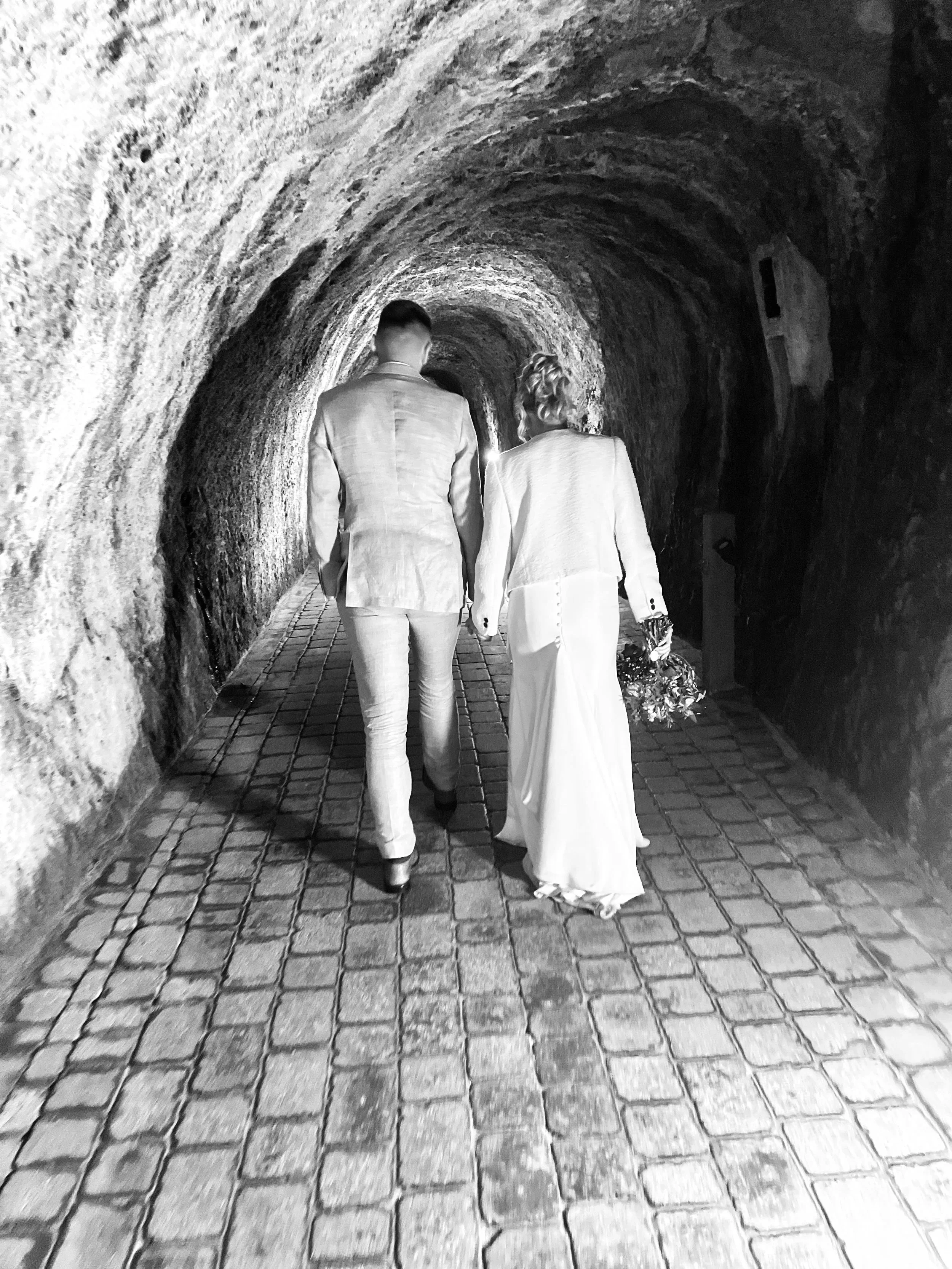 A couple in wedding attire walking hand in hand through a tunnel with stone walls and cobblestone floor.