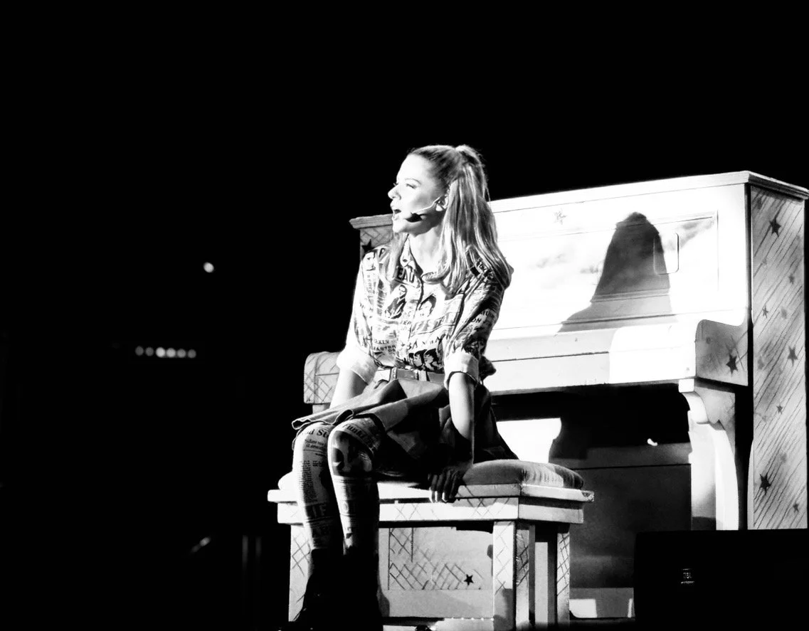A woman sitting on a bench on stage in front of a piano, wearing a patterned shirt and using a headset microphone, during a performance or speech.