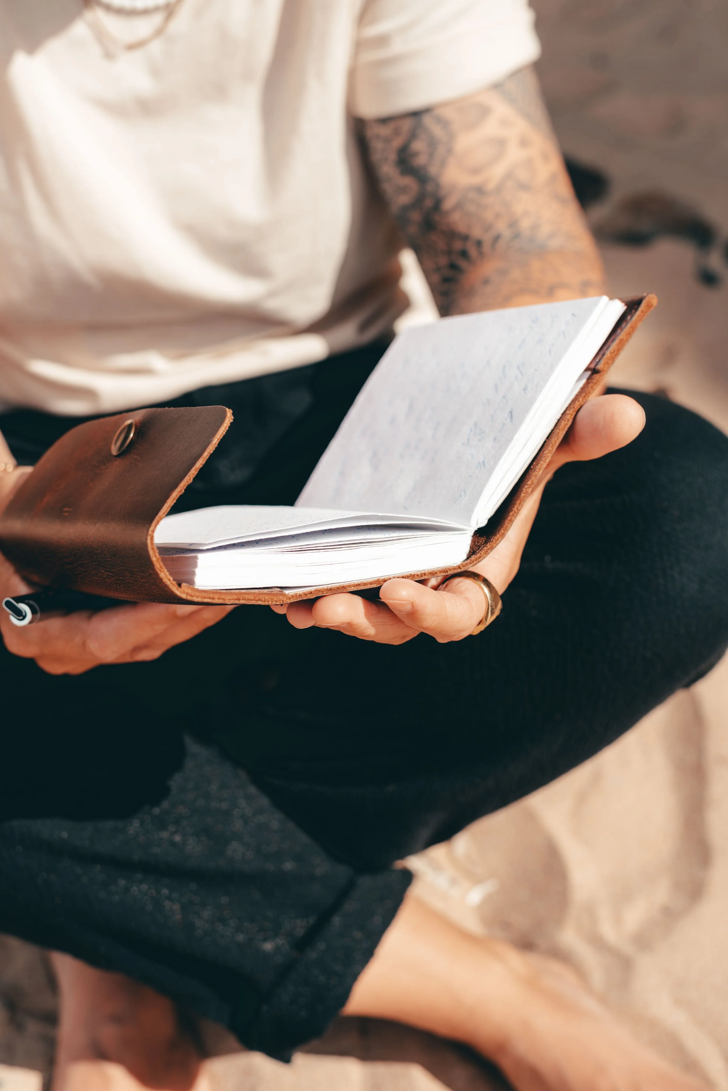 Person sitzt im Sand, liest in einem offenen Notizbuch mit braunem Lederumschlag.