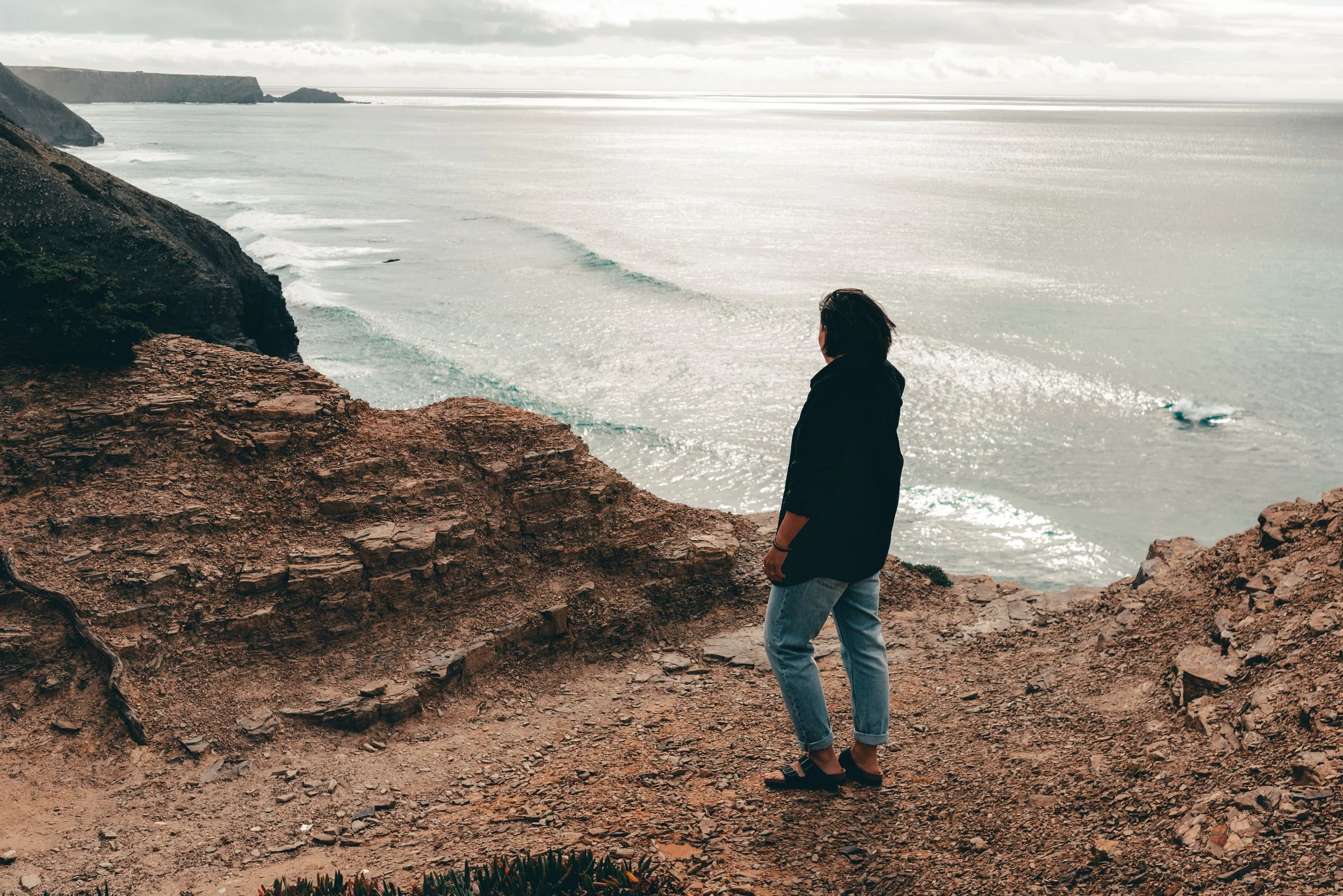 Eine Person steht an einer Klippe mit Blick auf das Meer, trägt dunkle Kleidung und Jeans, bei bewölktem Himmel.