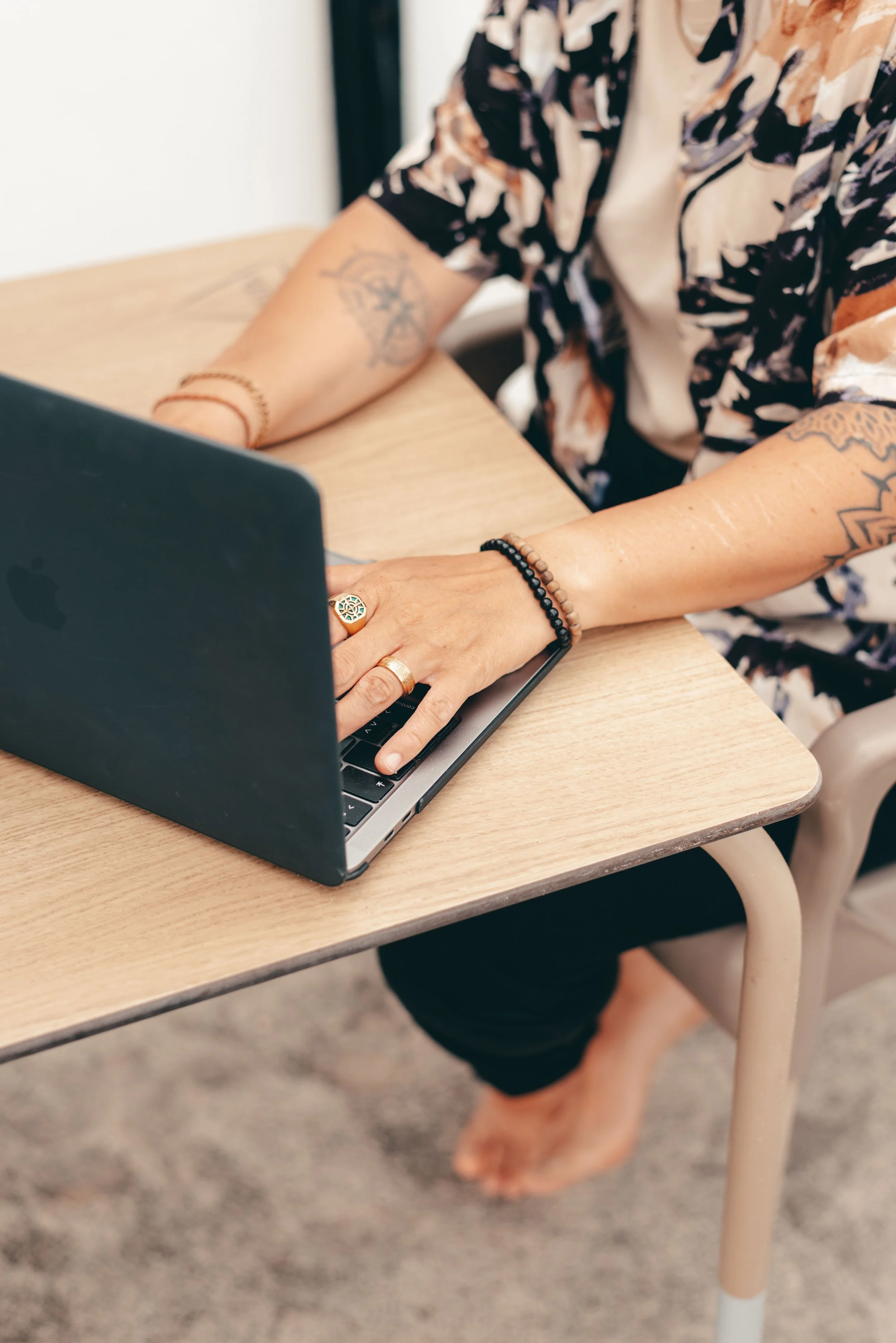 Eine Person tippt auf einem Laptop, während sie an einem Holztisch sitzt. Die Person trägt Ringe, Armbänder und hat Tattoos an den Armen. Die Person ist barfuß, mit einem schwarzen Rock oder Hose.