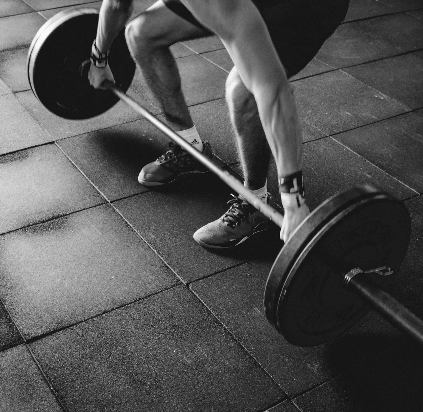 A person lifting a barbell with weights in a gym, wearing athletic shoes and shorts, on a rubber gym floor.