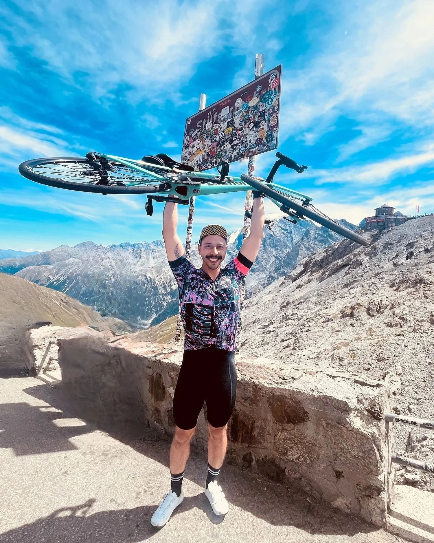 Reputable Physiotherapist Gianni Chng in cycling gear holding a bicycle above his head at the peak of the Stelvio Pass, Italy with a sign and mountains in the background.