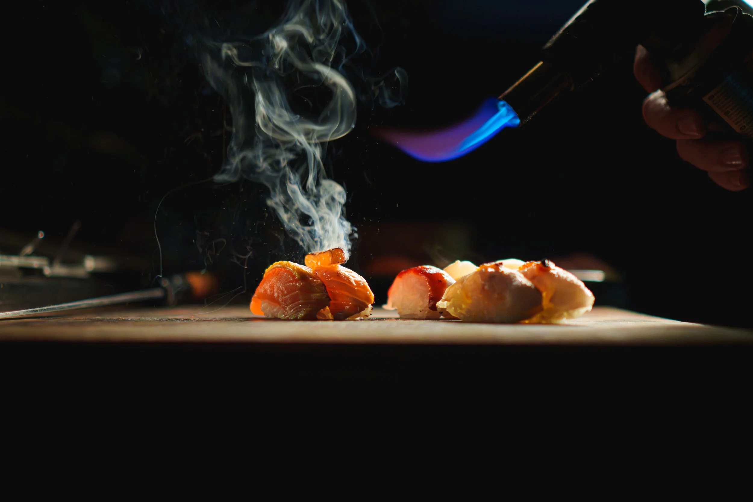 Sushibitar på en träskärbräda, med blåflamma från tändare som lägger till syre, ånga stiger från fisken.