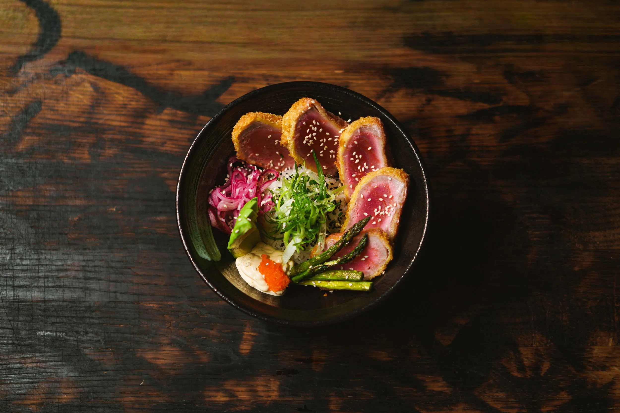 Tunnskuret, rå, rosa tonfisk med grönsaker och sesamfrön på ett svart porslin.