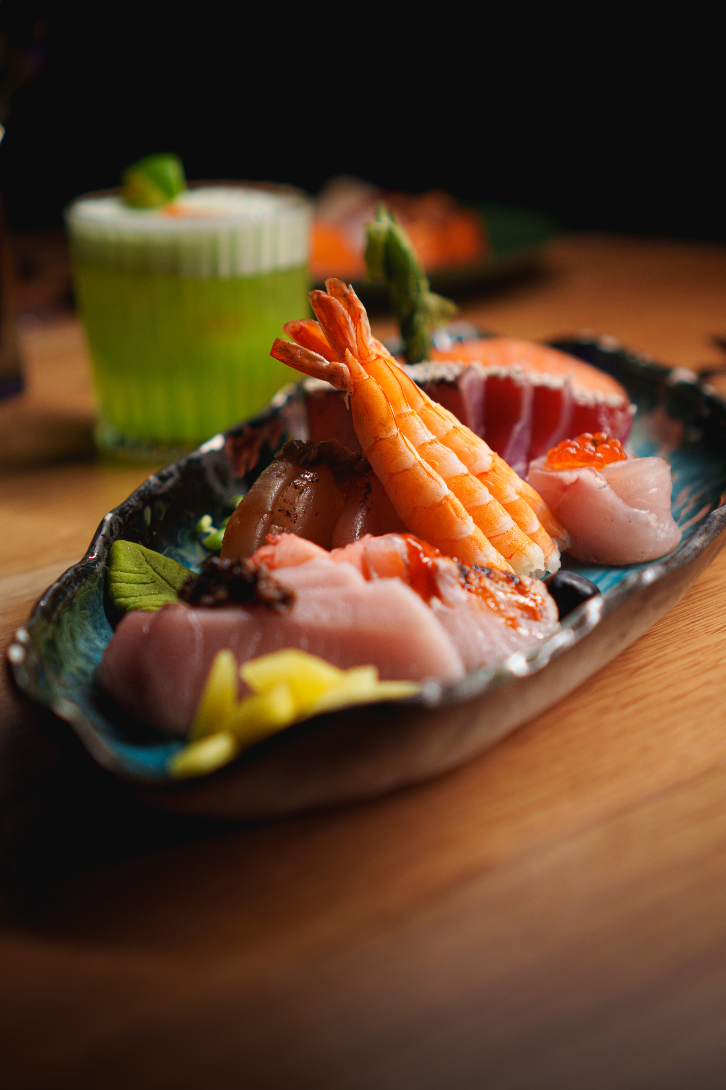 Färgglad japansk sashimi-tallrik med räkor, fisk och lax roe, serverad på en dekorativ tallrik, med en grön drink i bakgrunden.