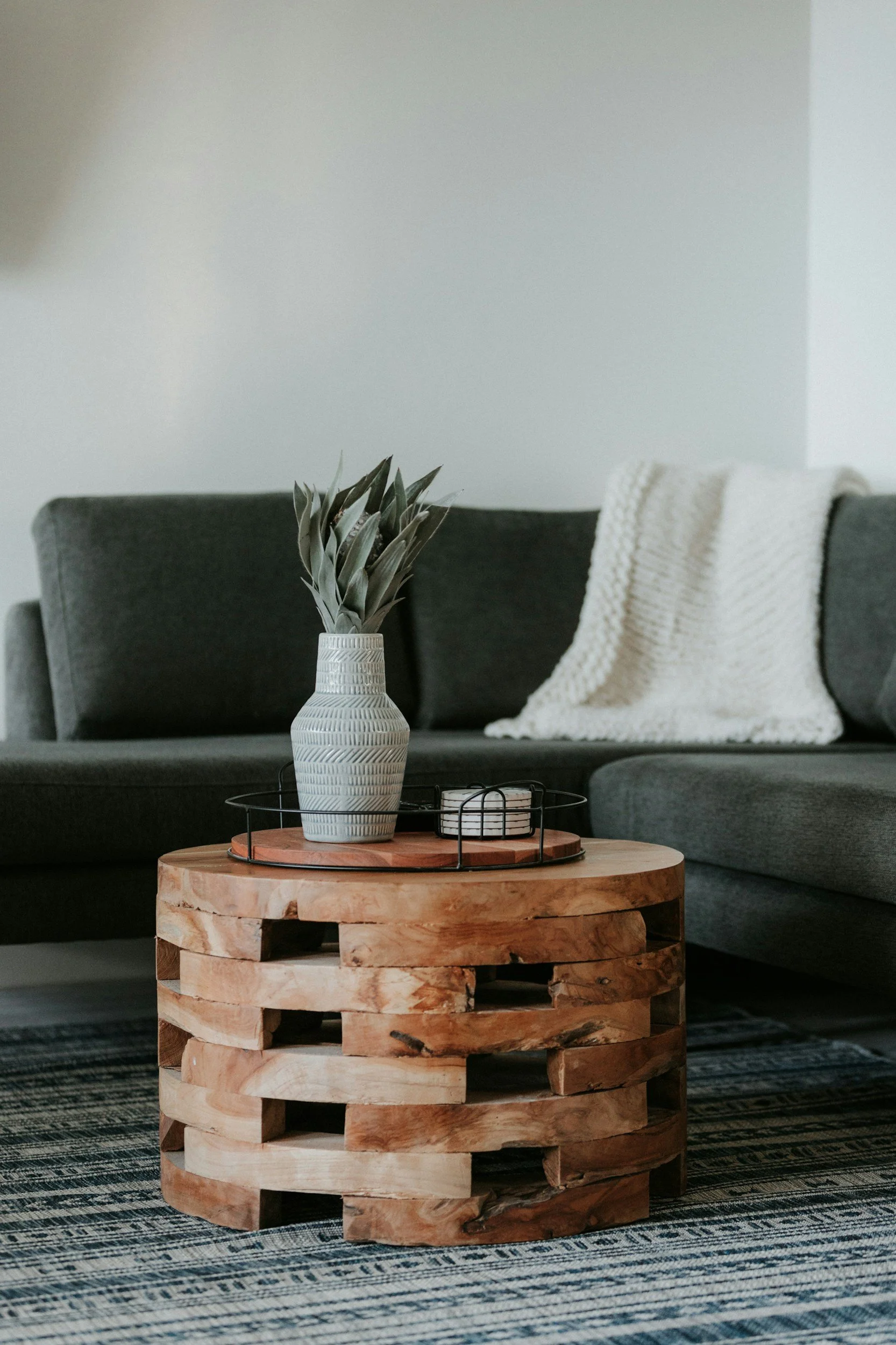 Living room with a round wooden coffee table, a gray sofa, a white knitted blanket, and a potted plant in a patterned vase.