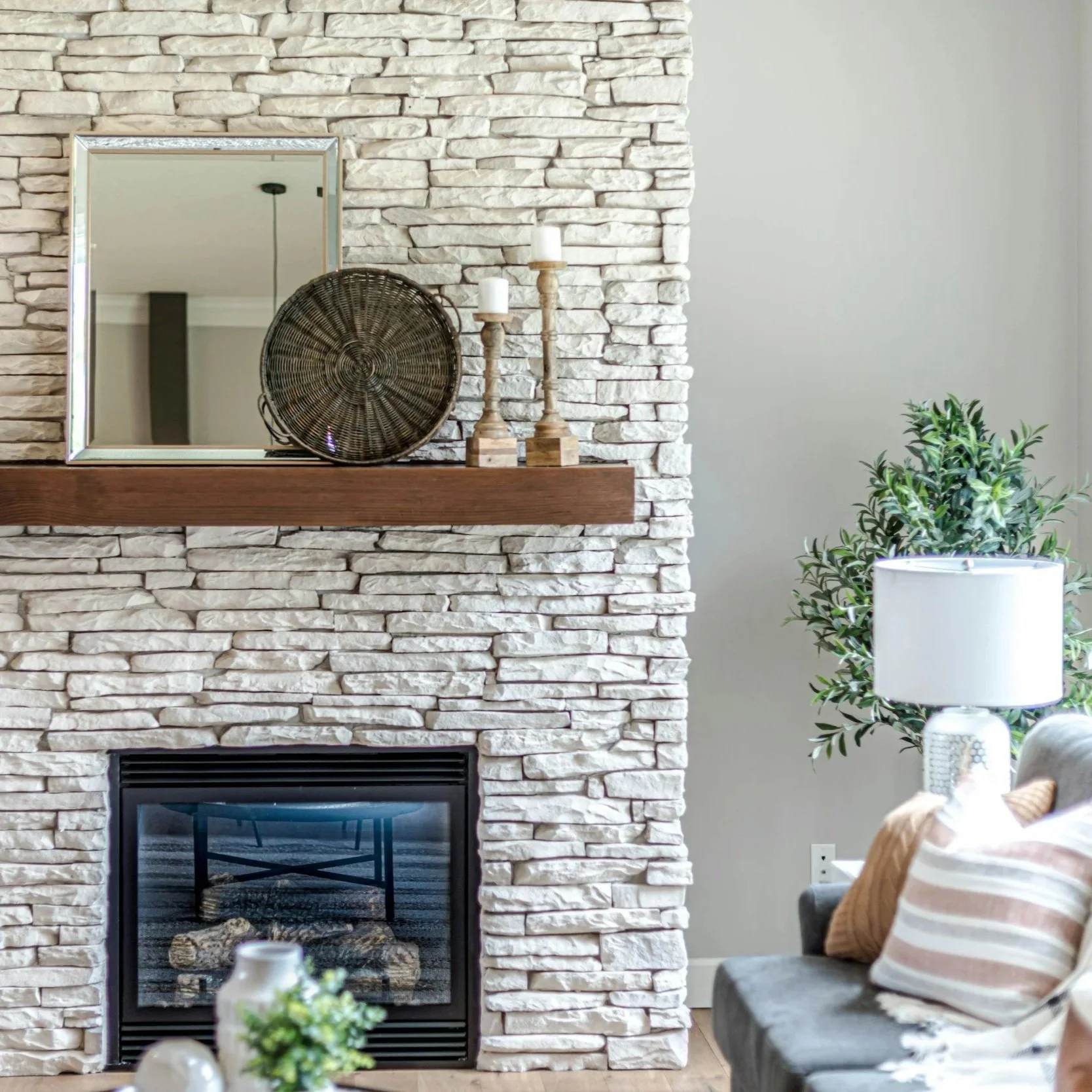 Living room with a stone fireplace, wooden mantel, mirror, wicker tray, two wooden candlesticks, a lamp, a green plant, and a couch with pillows.