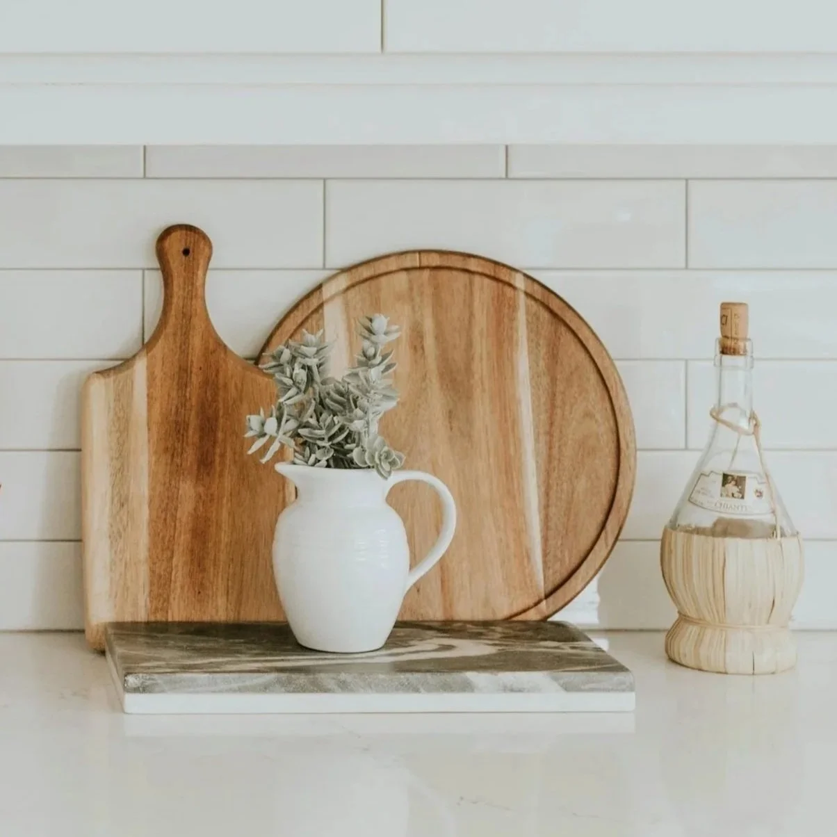 Decorative kitchen scene with wooden cutting boards, a white pitcher with greenery, and a glass bottle on a white countertop.