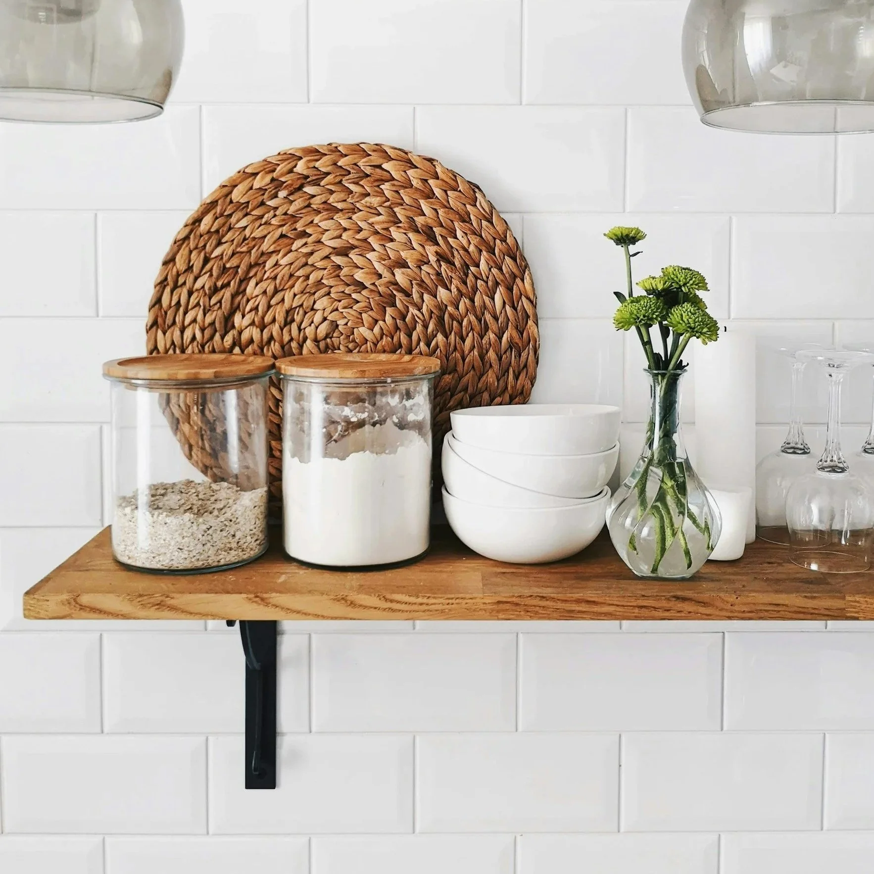 Kraft wooden shelf on a white tiled wall holding two glass jars with wood lids, white bowls, and a glass vase with green flowers.