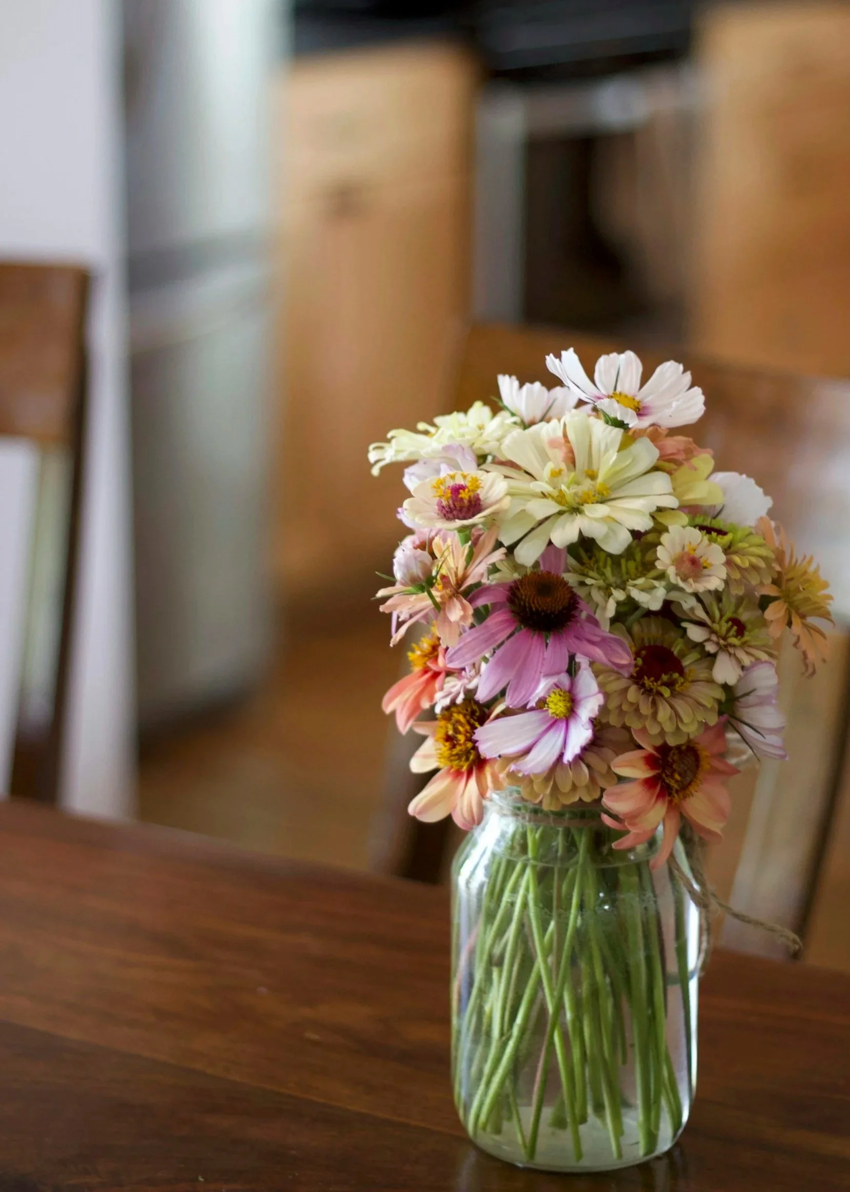 A glass jar filled with a bouquet of pink, white, and yellow flowers placed on a wooden table inside a room.