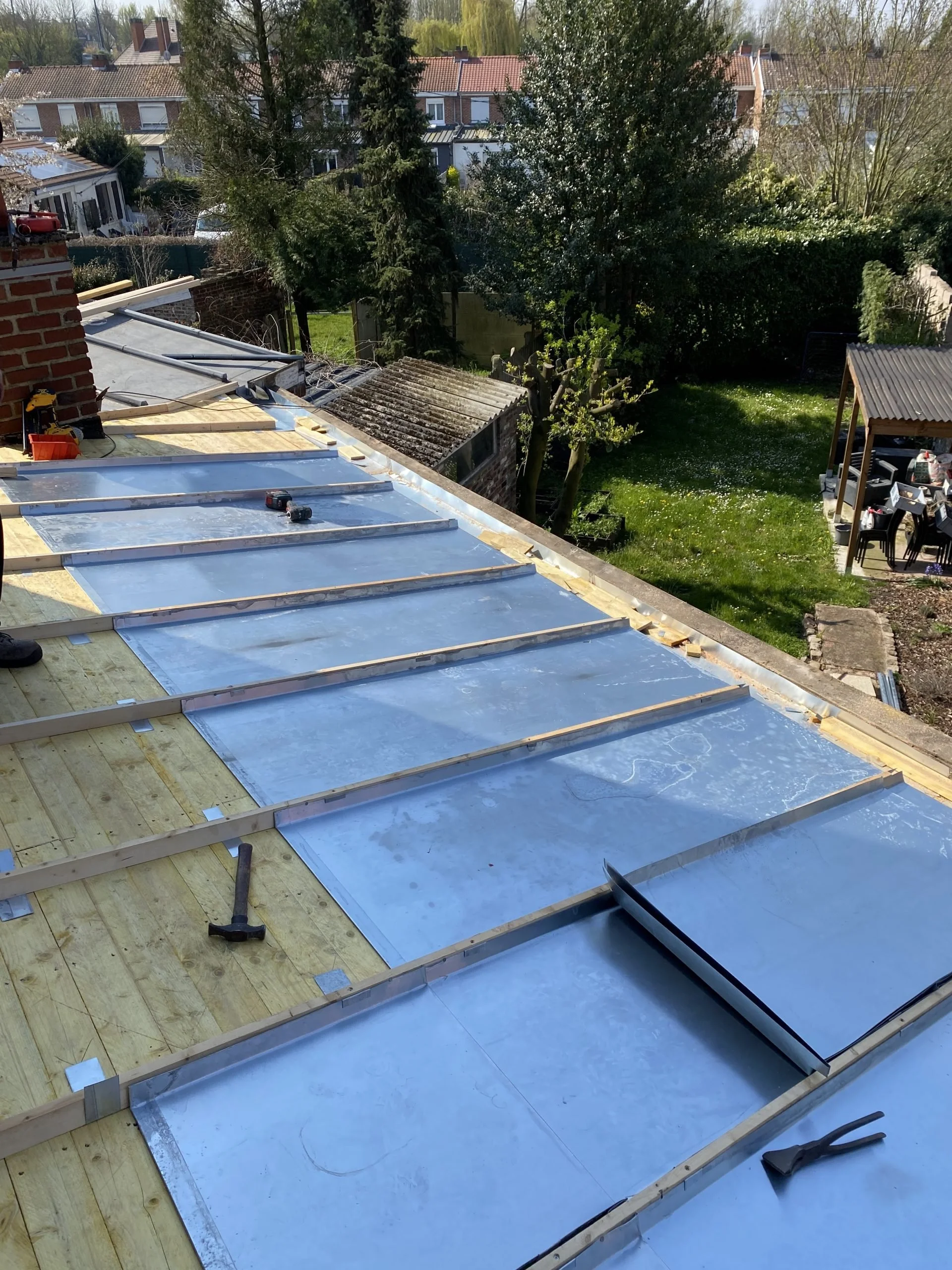 Construction d'une toiture avec des panneaux métalliques en cours, outils et bois visibles, vue sur un jardin et des maisons environnantes.