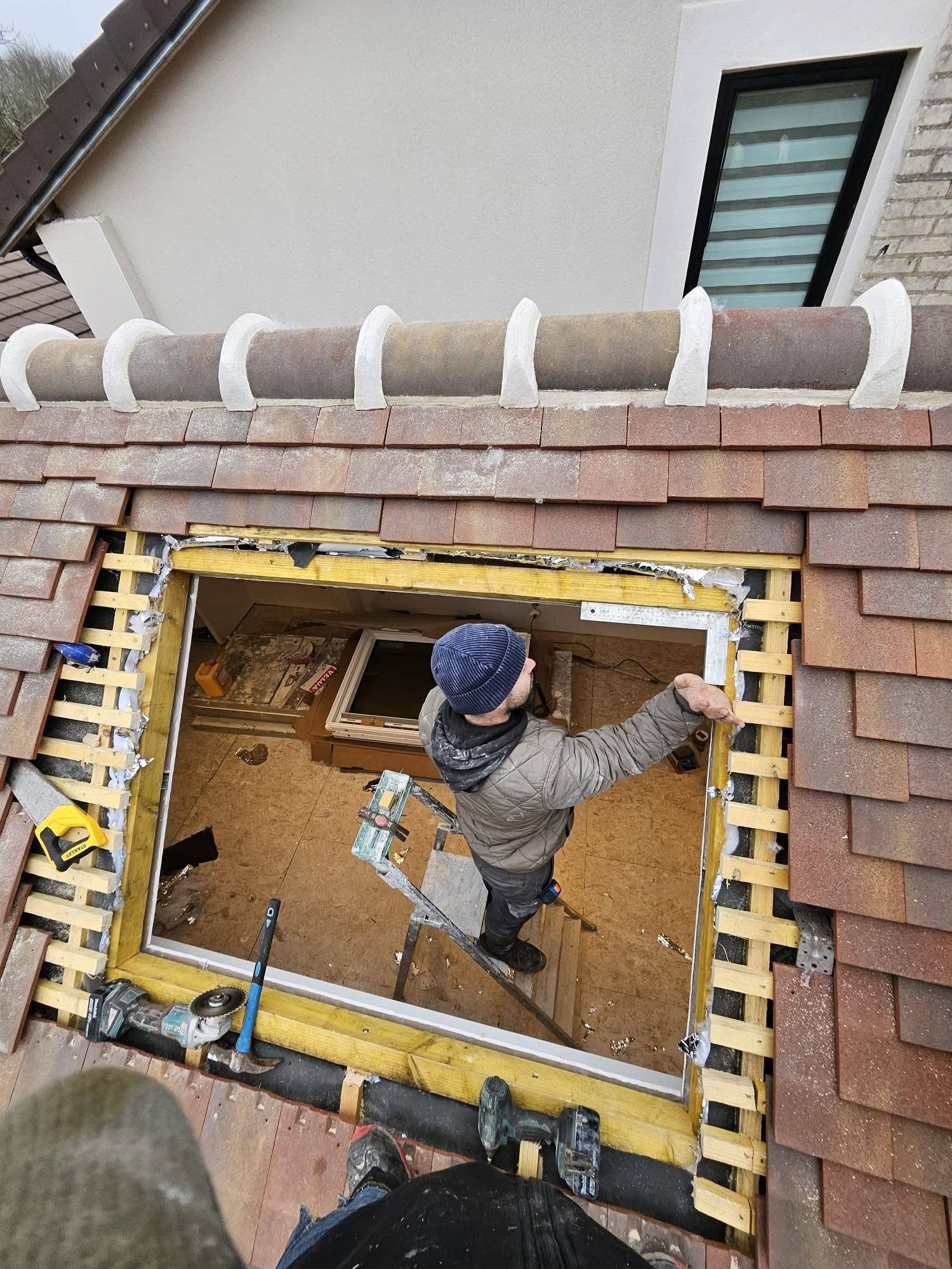 Photo prise d'en haut montrant un homme travaillant sur la rénovation d'une fenêtre de toit. Il se tient sur une échelle et entoure l'ouverture du carreau et de l'encadrement. Autour, le toit est en tuiles, et une partie du mur de la maison est visible.