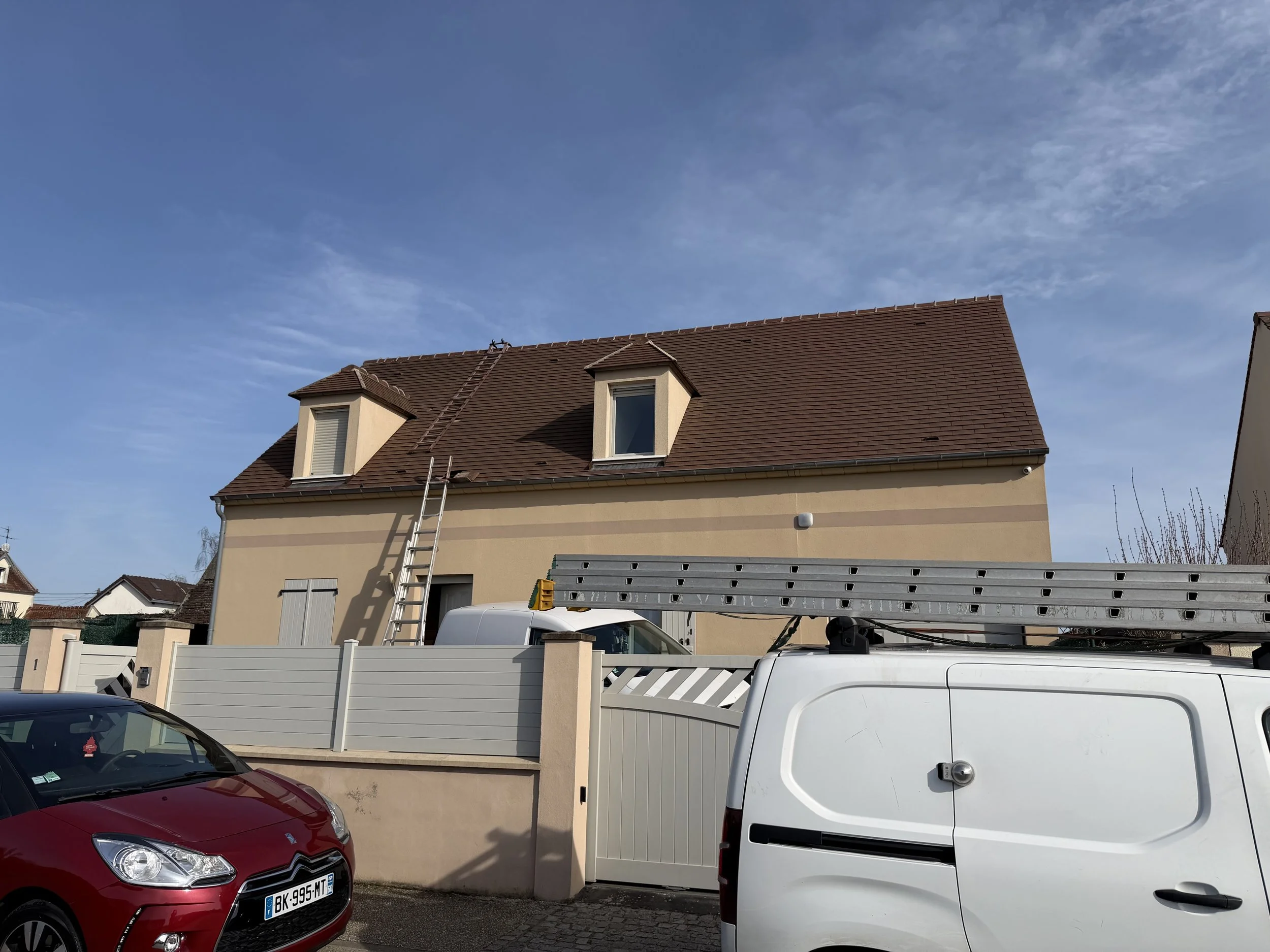 Une maison beige à deux étages avec des fenêtres à volets fermés et un toit en tuiles rouges. Deux fenêtres en pente sur le toit. Une échelle appuyée contre la maison. Deux véhicules garés devant la maison, une voiture rouge et une camionnette blanche avec une échelle sur le toit.