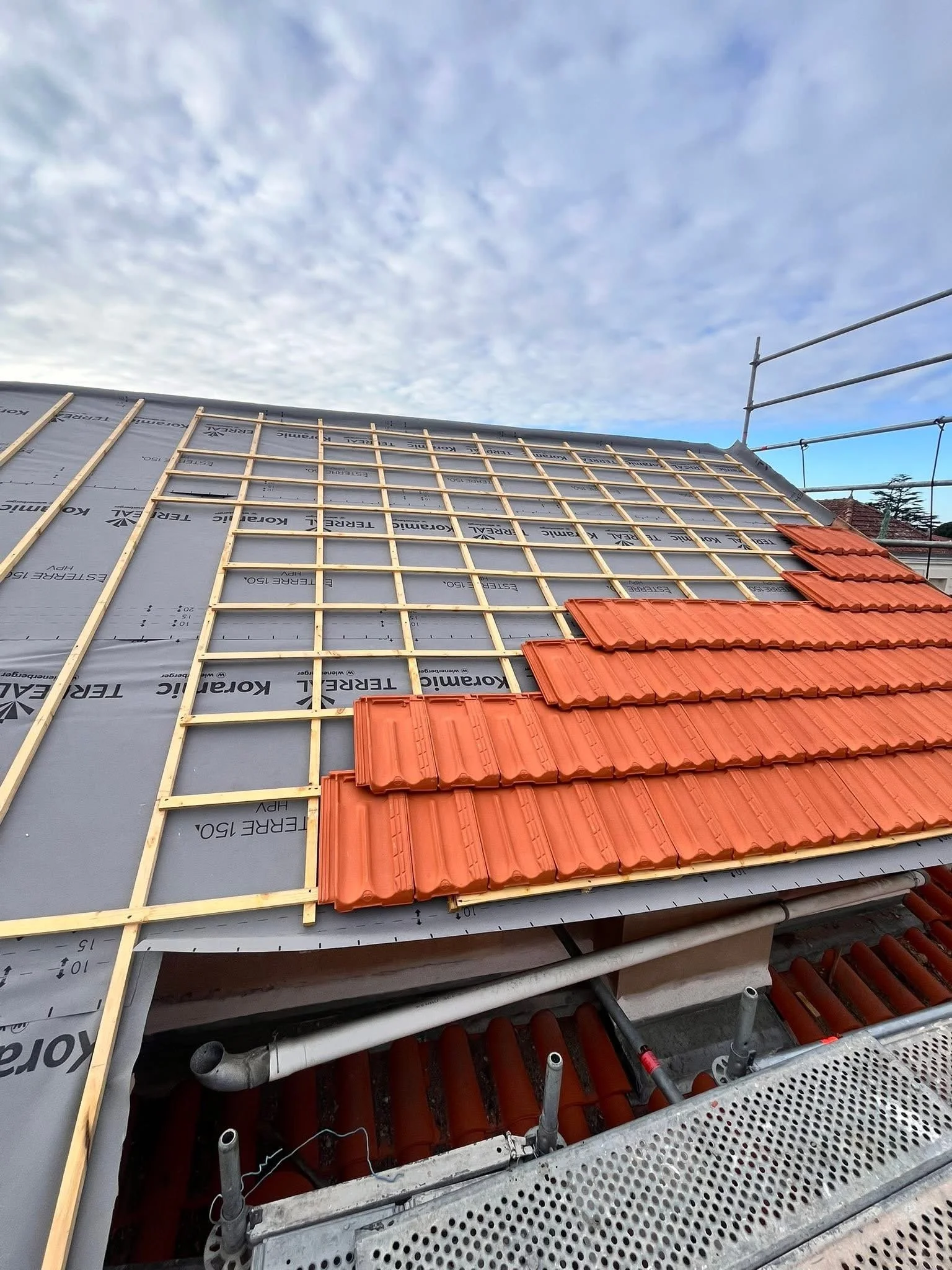 Construction en cours d'une toiture avec des tuiles rouges partiellement installées, avec une structure en bois et un ciel nuageux en arrière-plan.