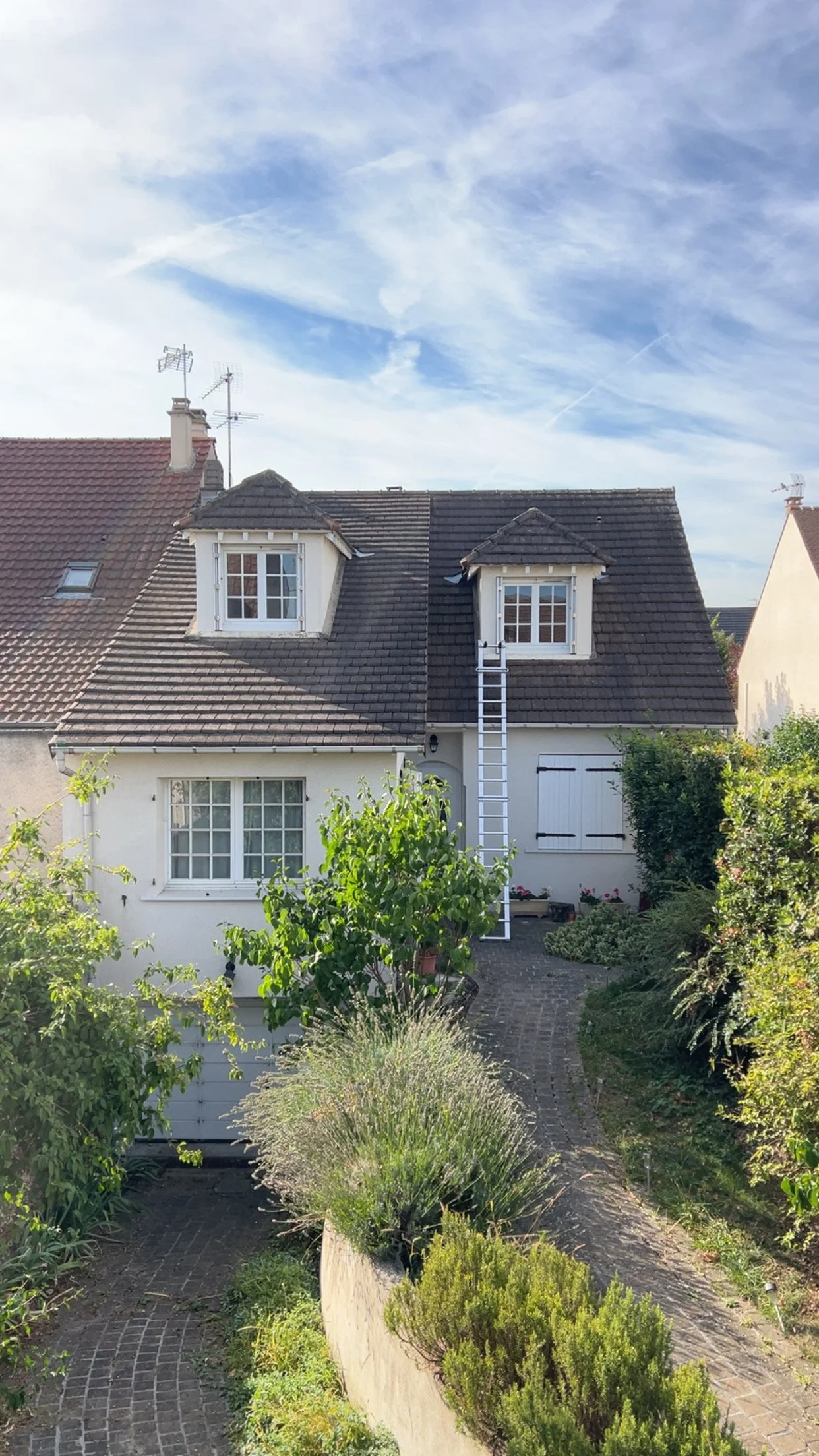 Maison blanche à deux étages avec un jardin fleuri, toiture en tuiles gris foncé, deux lucarnes, échelle blanche contre la façade et un ciel bleu avec quelques nuages.