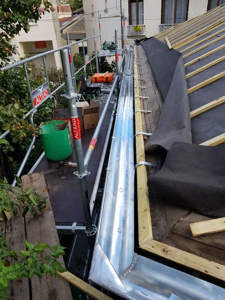 Toit en construction avec charpente en bois et une gouttière en zingue installée, entouré d'échafaudages.