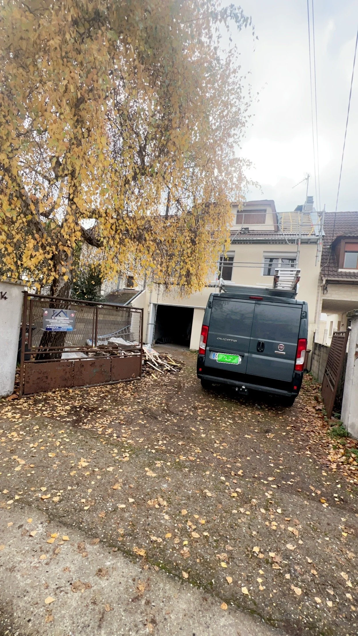 une cour extérieure avec un arbre à feuilles jaunes, une voiture noire, une maison et un portail en fer rouillé.