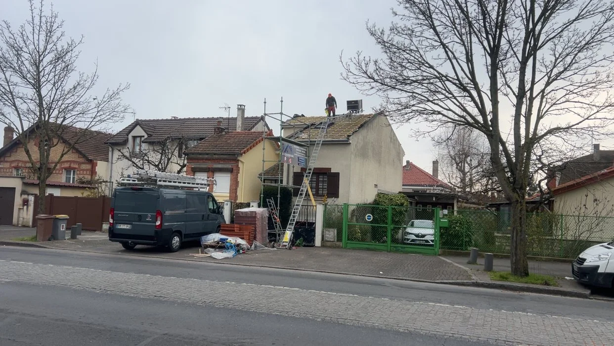 Travail de rénovation de toiture d'une maison avec un ouvrier sur le toit, échelle et matériaux de construction devant la maison, dans un quartier résidentiel.