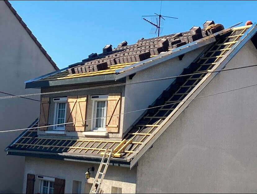 Une maison en cours de rénovation avec le toit en construction, des fenêtres à volets en bois et un échelle appuyée contre la façade.