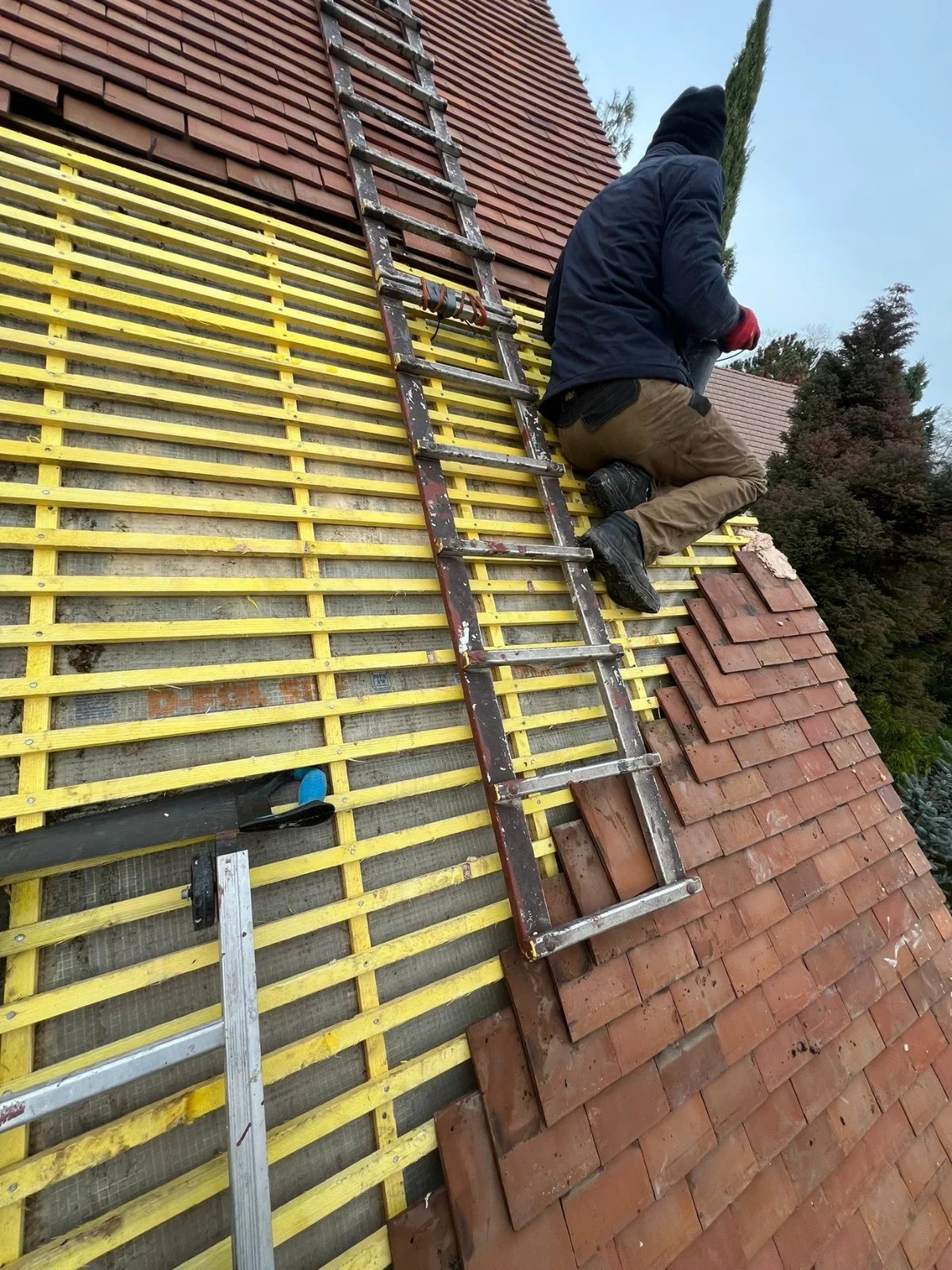 Un ouvrier en train de réparer ou de rénover le toit d'une maison, avec des lattes de bois jaunes et des tuiles en terre cuite.