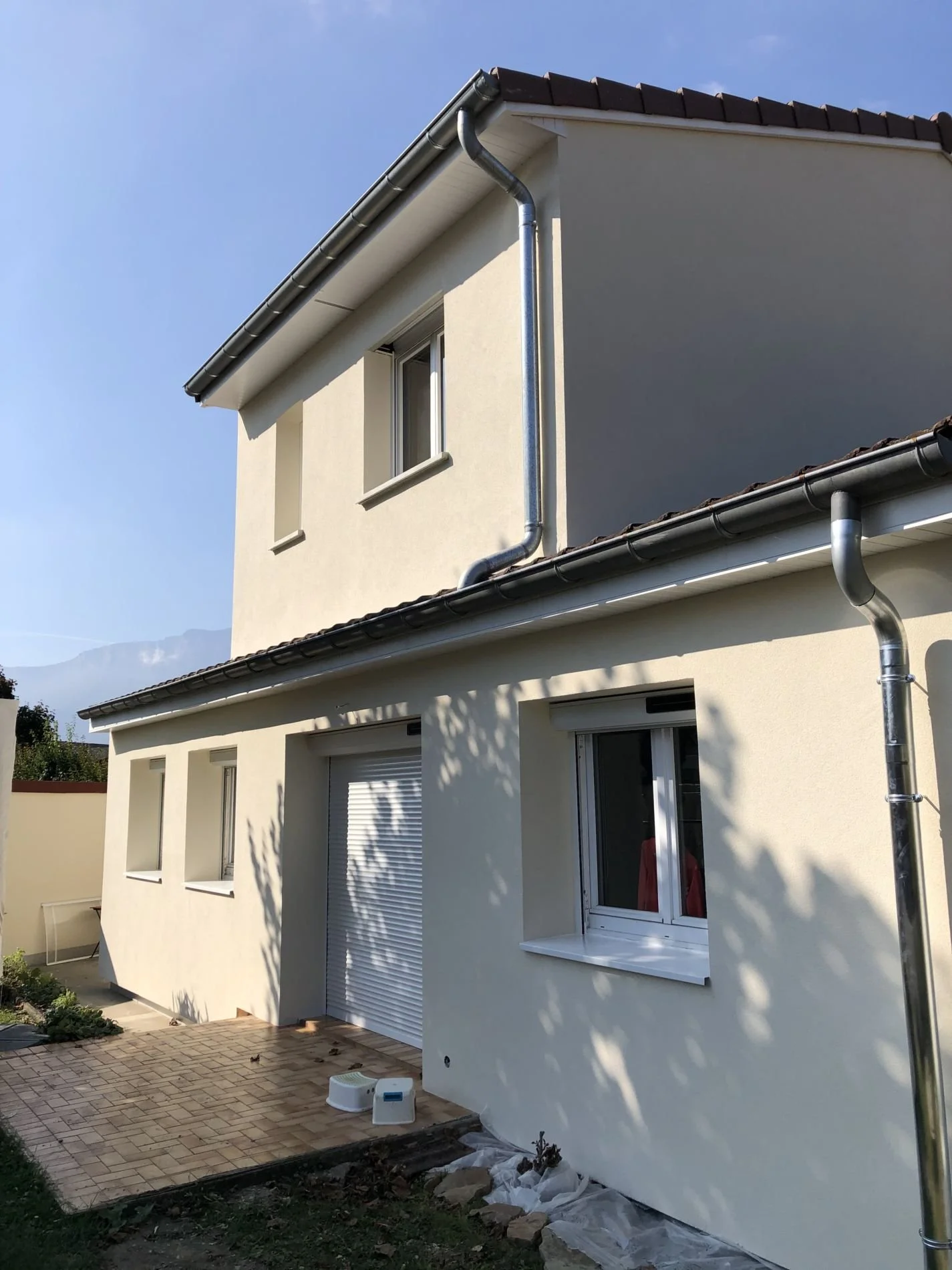 Maison à deux étages avec murs beige et tuiles brunes, fenêtres blanches, gouttières en métal, et un petit patio pavé en devanture.