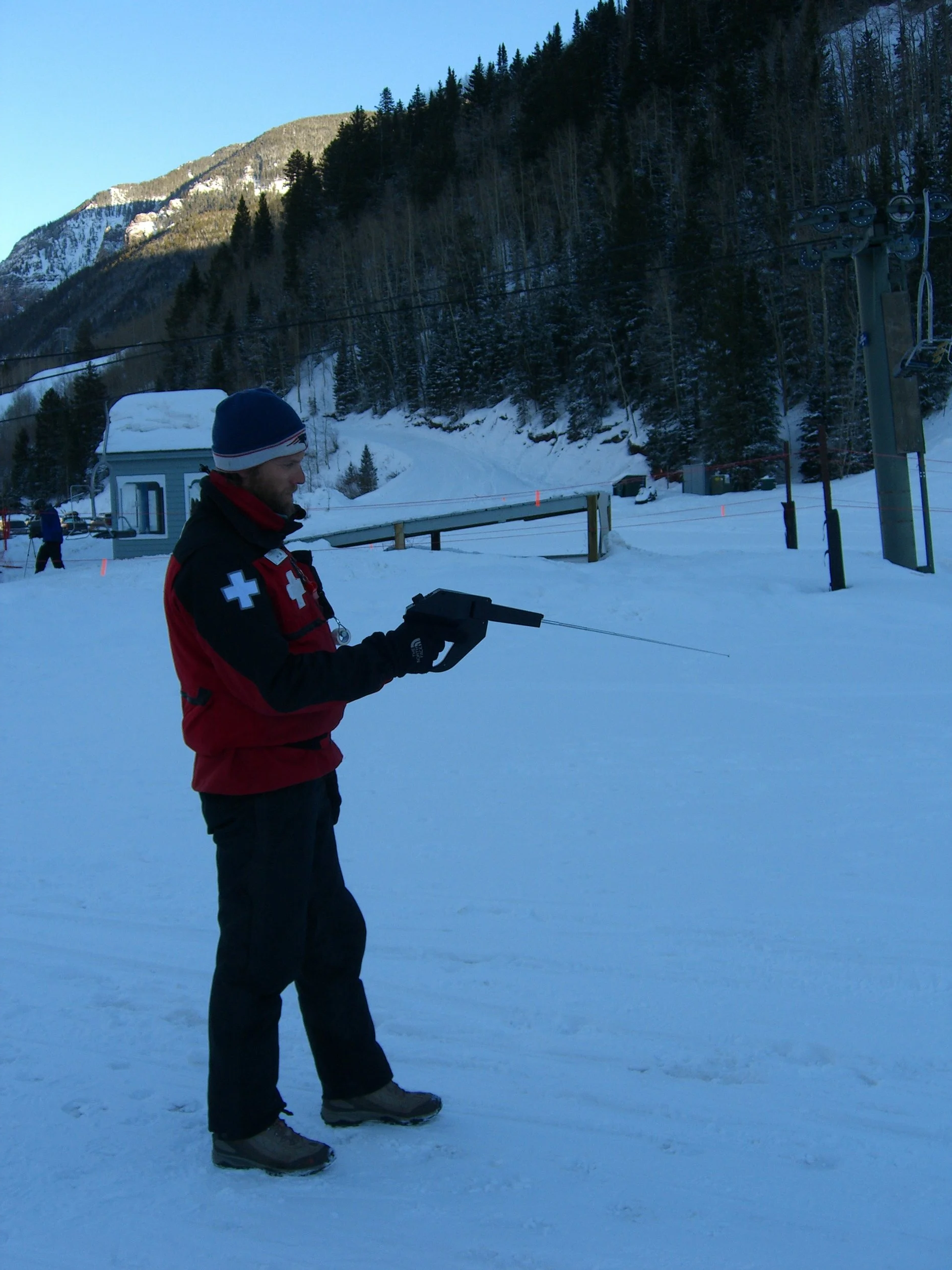A ski patrol member in red and black uniform holds a rescue sled, standing on a snowy ski slope with a mountain and pine trees in the background.