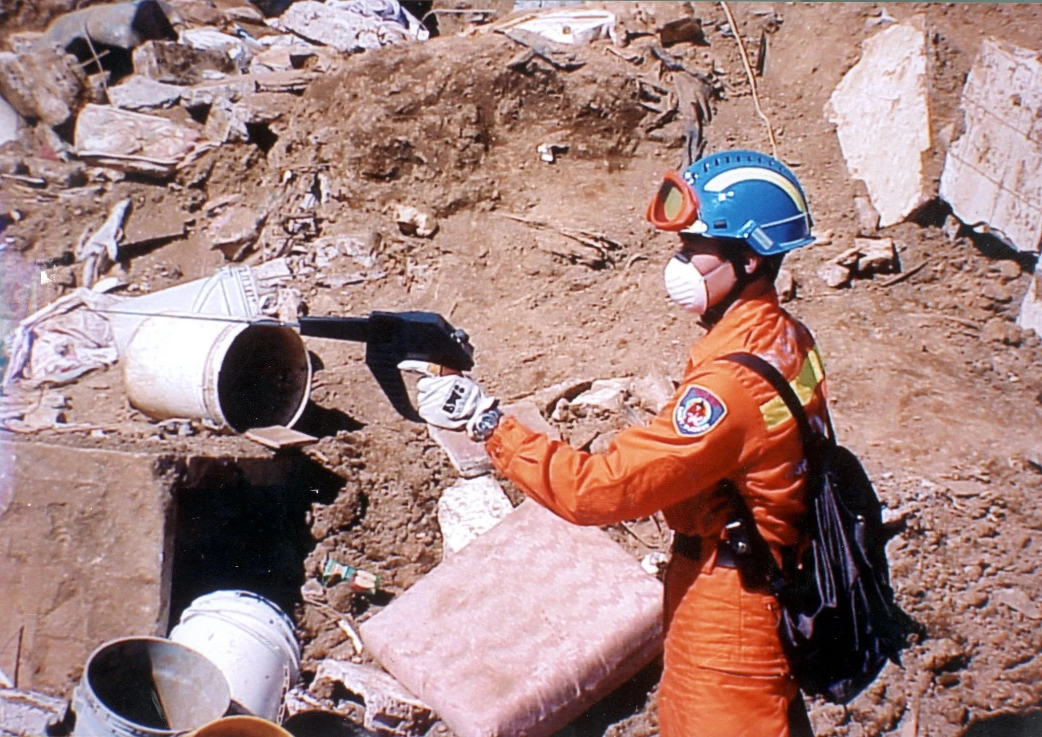 A rescue worker in orange uniform and helmet with face mask is working at a disaster site with rubble, rocks, and debris. The worker is holding a bucket or container with a tool.