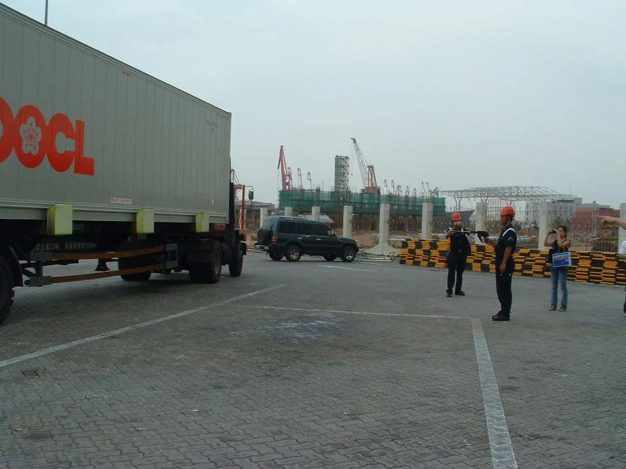 Construction site with cranes, workers in safety helmets, and vehicles including a truck and cars, on a paved parking area.
